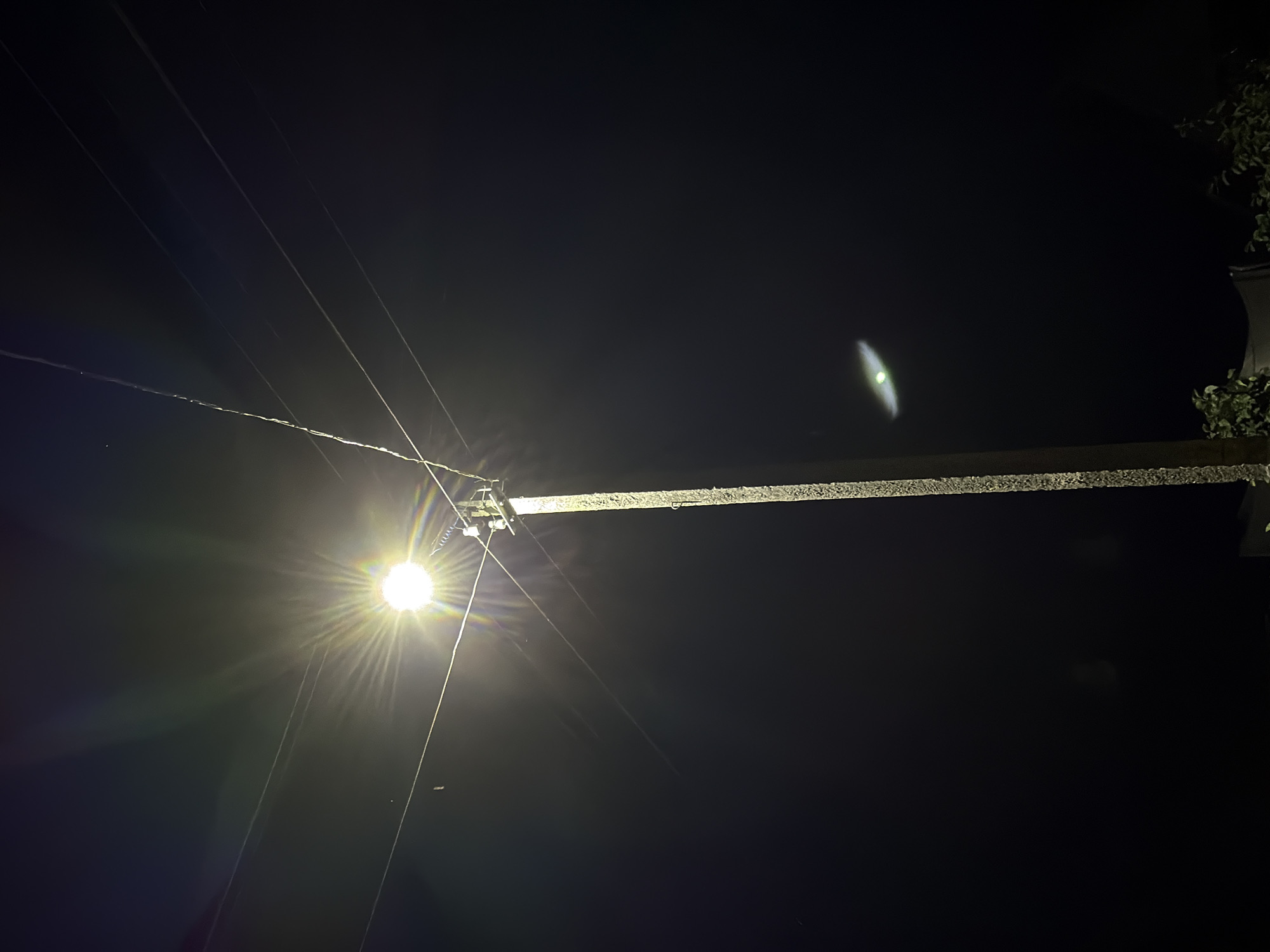 The image shows a street lamp at night in the Valyavichy village council, Belarus. The lamp is on, emitting a bright light that creates lens flares. Several wires are visible, suspended from the lamp post. The background is dark, indicating it is nighttime. There are some faint green lights in the distance, possibly from other lights or reflections. The bottom of the image shows a small amount of foliage, suggesting there may be trees or bushes nearby. The street lamp is made of concrete and has a textured surface. The overall scene is typical of a rural or suburban street at night.