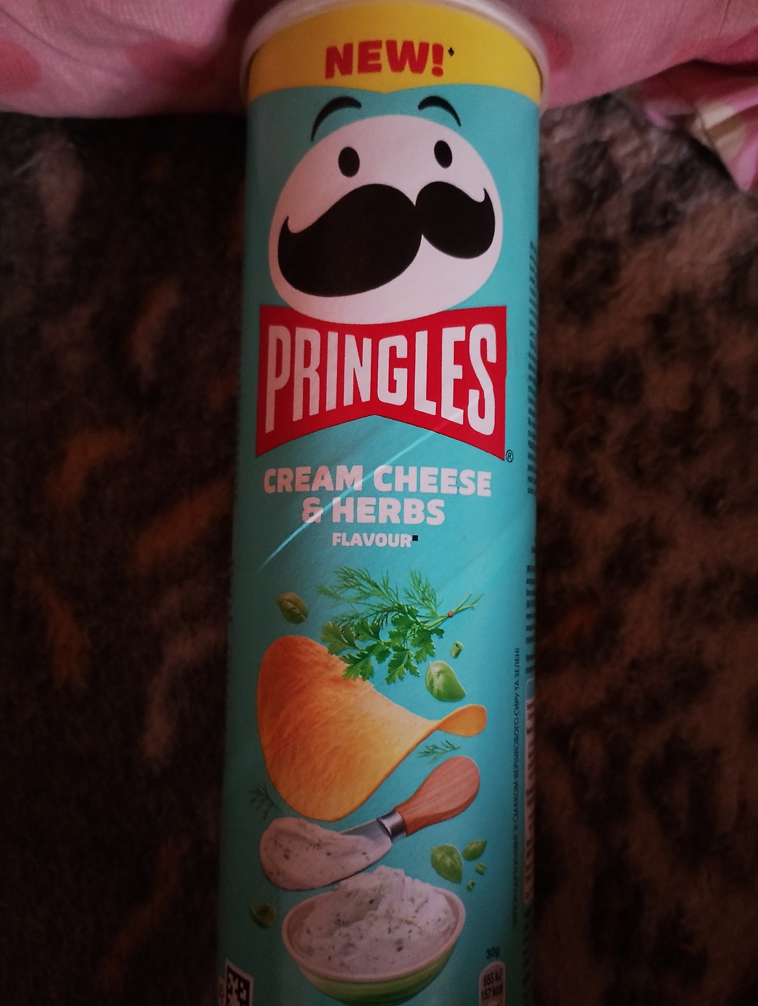 A Pringles can with a "New!" label is upright against a dark, textured surface, possibly a blanket or rug. The can is teal blue with a yellow rim and features the Pringles logo in red. Below the logo, it reads "Cream Cheese & Herbs" with "Flavour" underneath. An illustration of a potato chip, a sprig of dill, and a small bowl of dip are also depicted on the can. The top of the can has a pinkish fabric draped over it.
