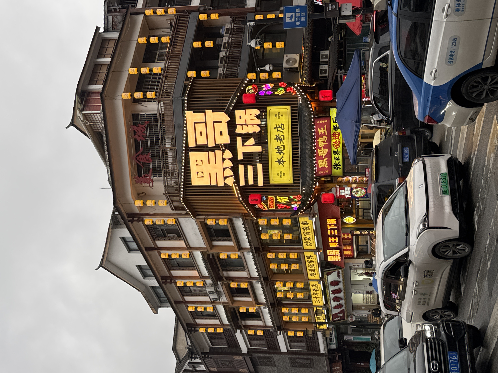 The image depicts a bustling commercial street scene in Wulingyuan, China, under an overcast sky. Multiple multi-story buildings line the street, featuring traditional Chinese architectural elements such as curved eaves and balconies with railings. The buildings are primarily light-colored with dark wood accents and are extensively adorned with rows of illuminated orange-yellow square lanterns.
The central building features a prominent, vertically oriented yellow neon sign displaying large Chinese characters "黑哥三下锅" (Hēi Gē Sān Xià Guō), with a smaller yellow sign below reading "本地老店" (Local Old Store). This building also displays a red sign on a second-floor balcony, multiple air conditioning units, and security cameras. Below the main sign, a neon duck character with purple and red lights is visible.
To the left, adjacent buildings display other illuminated storefront signs, including "三十年老店" (Thirty-Year Old Store), "地道张家界" (Authentic Zhangjiajie), and "黑哥鸭王" (Black Brother Duck King) with "张家界必吃店" (Zhangjiajie Must-Eat Restaurant) beneath it. A blue road sign indicating "电子警察" (Electronic Police) is mounted on a pole.
The street is paved and appears wet or reflective, indicating a recent rain or damp conditions. Multiple vehicles are present in traffic. In the foreground, a black SUV (license plate 湘G 01761) is on the left. A white multi-purpose vehicle (MPV) with "滴滴" (Didi) branding (license plate 湘G D18319) occupies the center. A blue and white taxi with "投诉电话: 12345" (Complaint Hotline: 12345) on its side is visible on the right. Additional vehicles, including a black SUV, are further back. A dark blue canopy or umbrella is positioned on the sidewalk or near the road's edge. Few individuals are discernible on the sidewalks or near the storefronts, their actions indistinct.