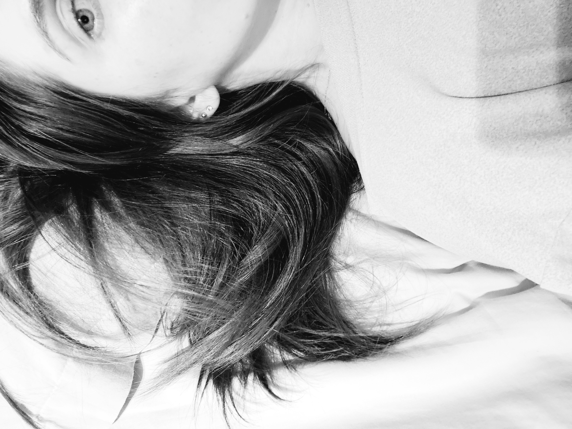 A black and white image shows part of a person's face and head lying on a bed. The visible part of the face includes an eye, part of the cheek, and an ear with multiple piercings. Dark hair is spread across the bed around the person's head. The person appears to be wearing a light-colored garment. The scene is indoors, implied to be in a bed in a bedroom, and the city location, Chaikovsky, Russia, is only provided as context but cannot be determined from the image itself.
