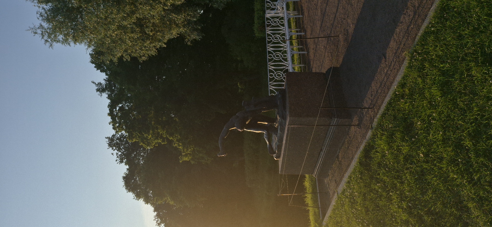 This media file depicts a scene in a park, likely during the late afternoon or early evening given the long shadows cast by the setting sun. The image is oriented vertically, with the sky taking up the left portion and a grassy area and pathway on the right.
The main subject is a bronze statue of two figures, seemingly engaged in an athletic activity. One figure appears to be in motion, perhaps reaching out or running, while the other is partially obscured, possibly reacting or participating. The statue is mounted on a dark, rectangular pedestal, which in turn sits on a paved area. Adjacent to this paved area is a neatly trimmed grassy lawn, dotted with small white flowers.
In the background, a white ornate fence or railing runs along the edge of the paved area. Behind the fence, trees form a dense, dark green canopy, suggesting a mature park or garden setting. The sunlight casts a warm, golden glow, illuminating the grass and the right side of the statue, while leaving the left side in shadow.
The overall impression is one of tranquility and perhaps historical significance, typical of a public park in a place like Pushkin, Russia, known for its imperial palaces and gardens. There is no visible text in the image.
