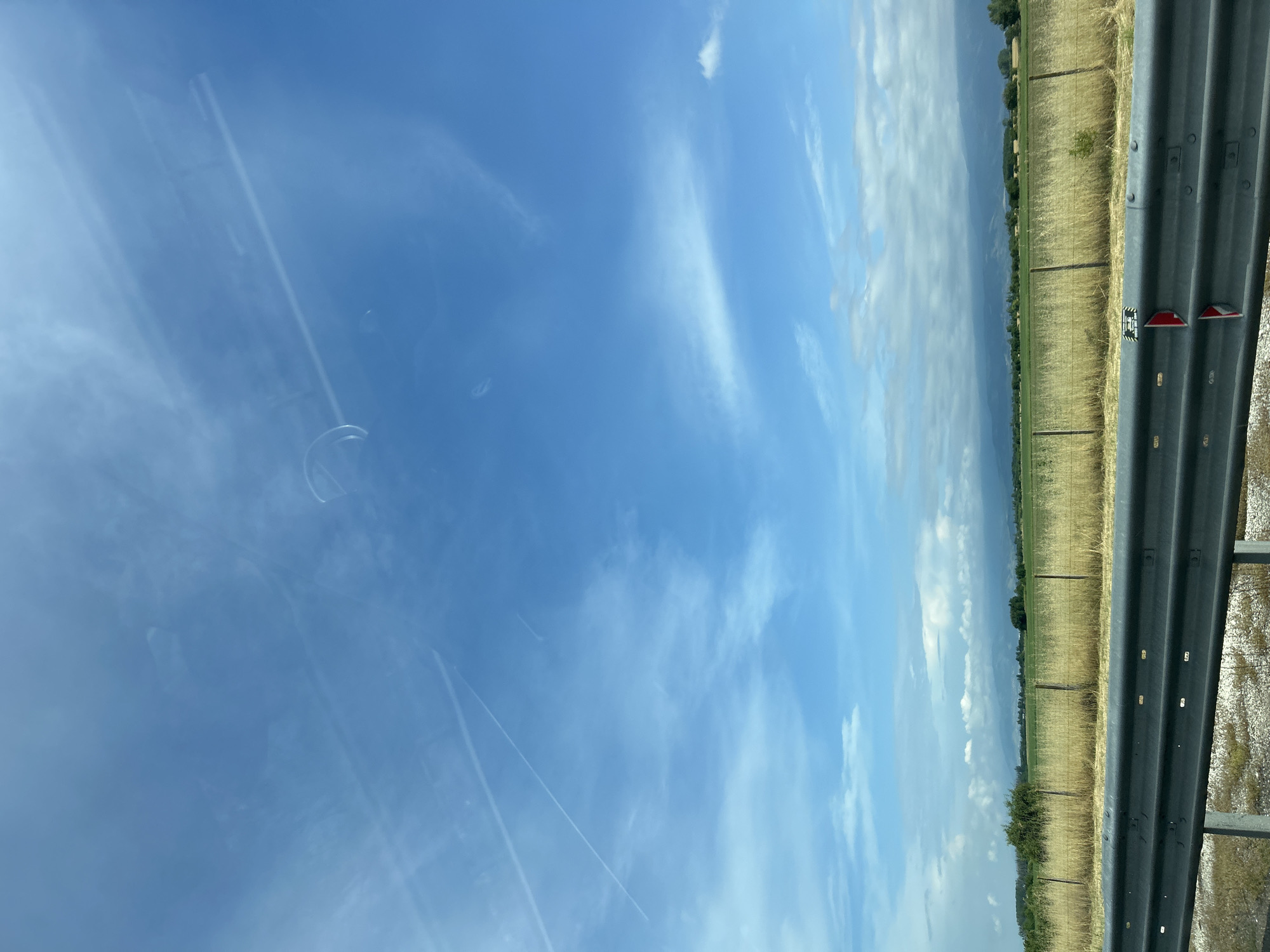 This media file captures a sunny daytime landscape from the perspective of a moving vehicle, likely in Malo Konare, Bulgaria. The image, originally rotated, shows a wide-angle view dominated by a clear blue sky with scattered white, wispy clouds and several faint airplane contrails. Prominent glare and reflections on what appears to be a car window are visible across the sky, including faint circular and linear patterns.
In the right foreground, a dark grey, corrugated metal guardrail runs horizontally across the frame, indicating a road or highway. Attached to the guardrail is a small white rectangular marker with black text, partially visible as "T 200" and possibly "196" or "1967" below it. Below this, two red reflective strips are also affixed to the guardrail.
Beyond the guardrail, a dry, golden-brown field stretches into the midground, traversed by a line of fence posts. In the distance, a hazy horizon line reveals a band of green trees and distant hills or mountains. The weather appears bright and clear, characteristic of a sunny day, possibly during the summer or a dry season given the color of the fields. The scene conveys a sense of travel through a rural, open landscape. No individuals are visible in the image.