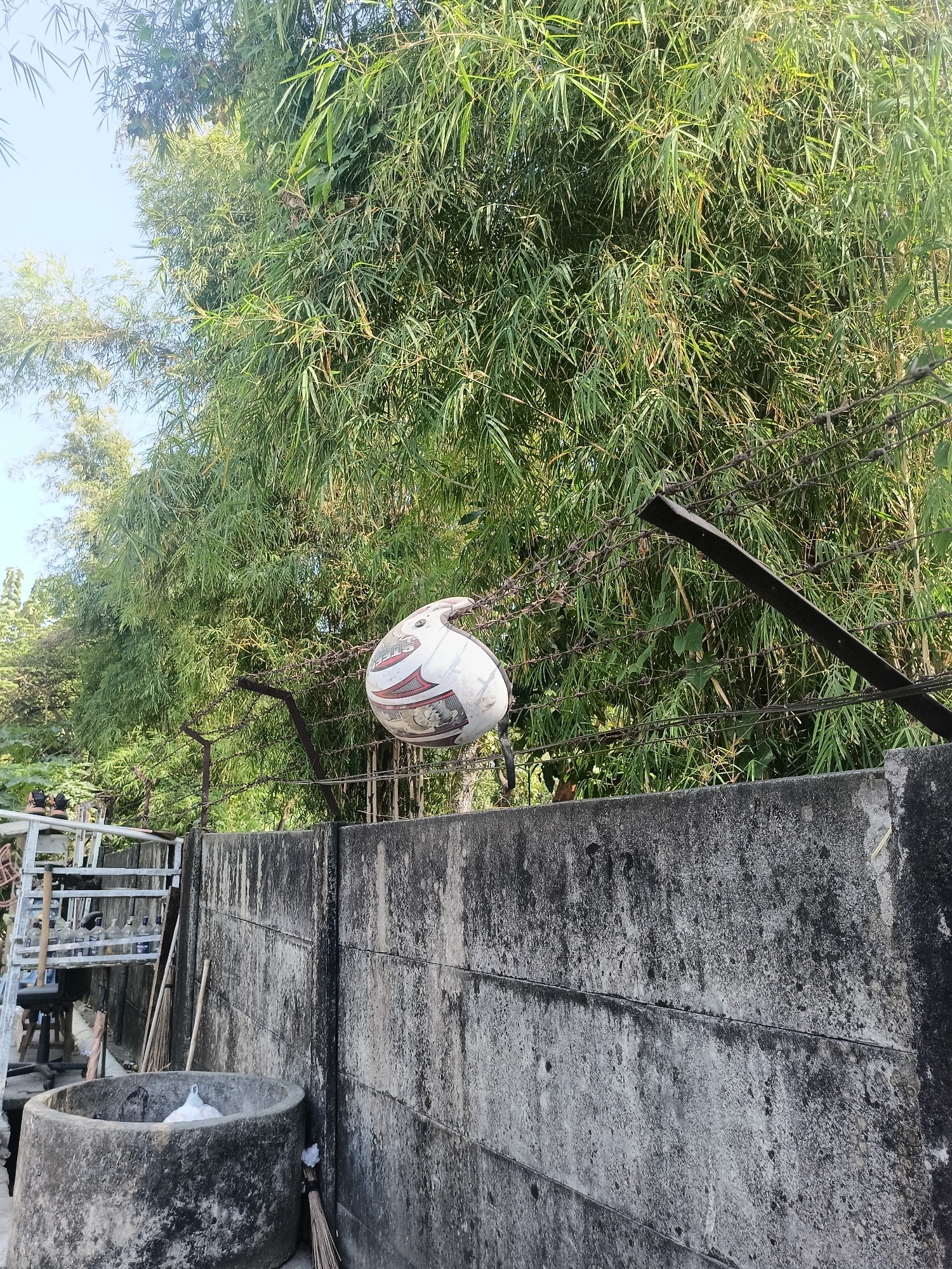 A white motorcycle helmet with red and black markings is impaled on barbed wire, suspended above a concrete wall. The helmet is positioned in front of a dense bamboo forest. The concrete wall runs horizontally across the lower portion of the image and is weathered and stained. To the left of the wall, there is a circular concrete structure with a white object inside. A metal ladder and some tools are also visible on the left side. The scene appears to be in an outdoor area. The location is identified as Gedangan, Indonesia.