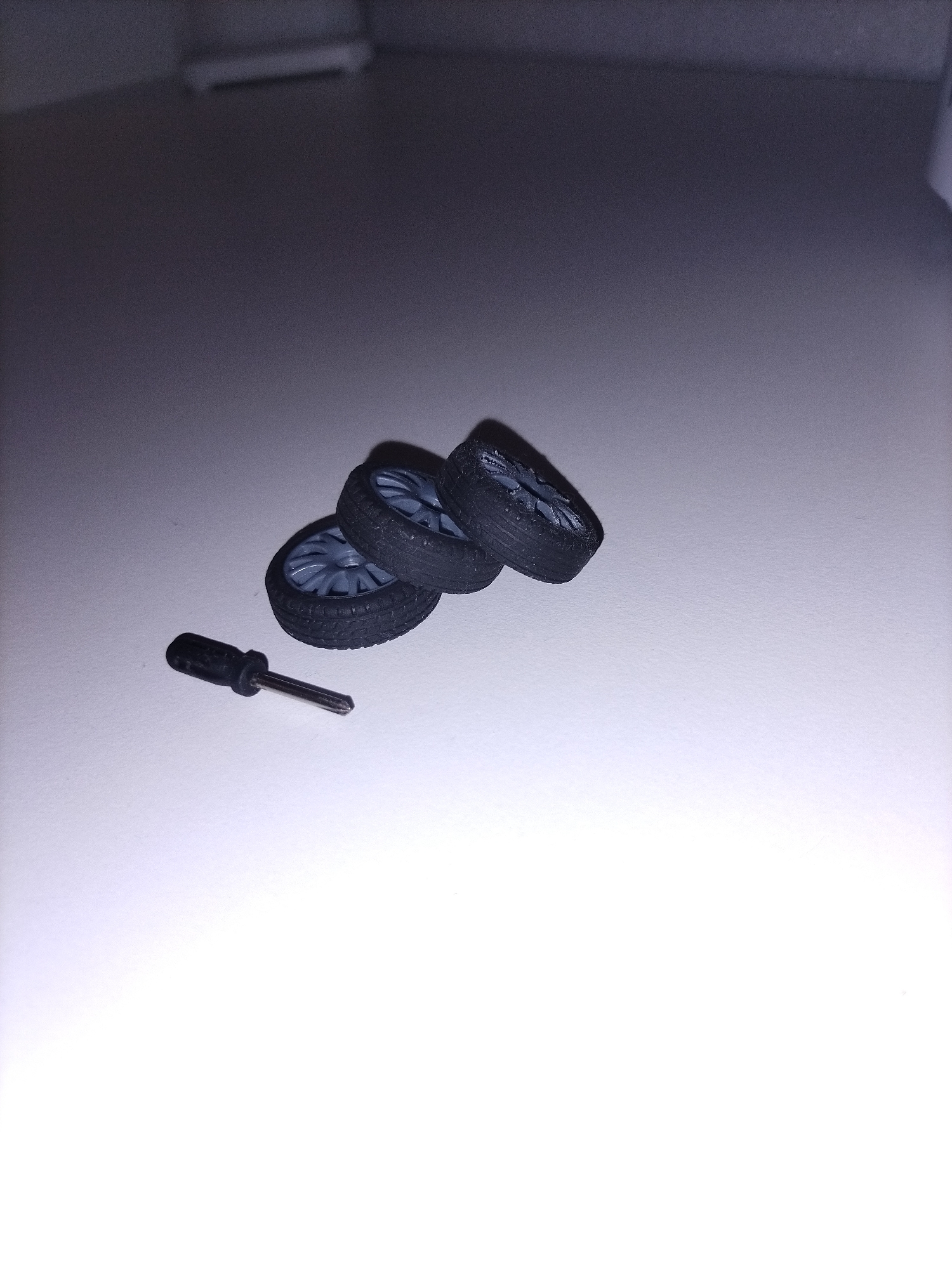 The image displays an indoor scene with three miniature toy car wheels and a small screwdriver resting on a light-colored, flat surface, likely a table or desk. The toy wheels feature black rubber tires and light grey, multi-spoke rims, positioned closely together towards the upper right of the frame. Two wheels lie on their sides, while one is slightly tilted. To their left, a small screwdriver with a black handle and a silver, Phillips-head shaft lies horizontally.
A light source from the upper left creates soft shadows cast by the objects onto the surface. The background is out of focus, appearing as a plain, darker grey wall or room partition. No people are visible, and no specific activity is taking place; the objects are static. There is no visible text in the image. The time of day and external weather conditions cannot be determined from this indoor setting.