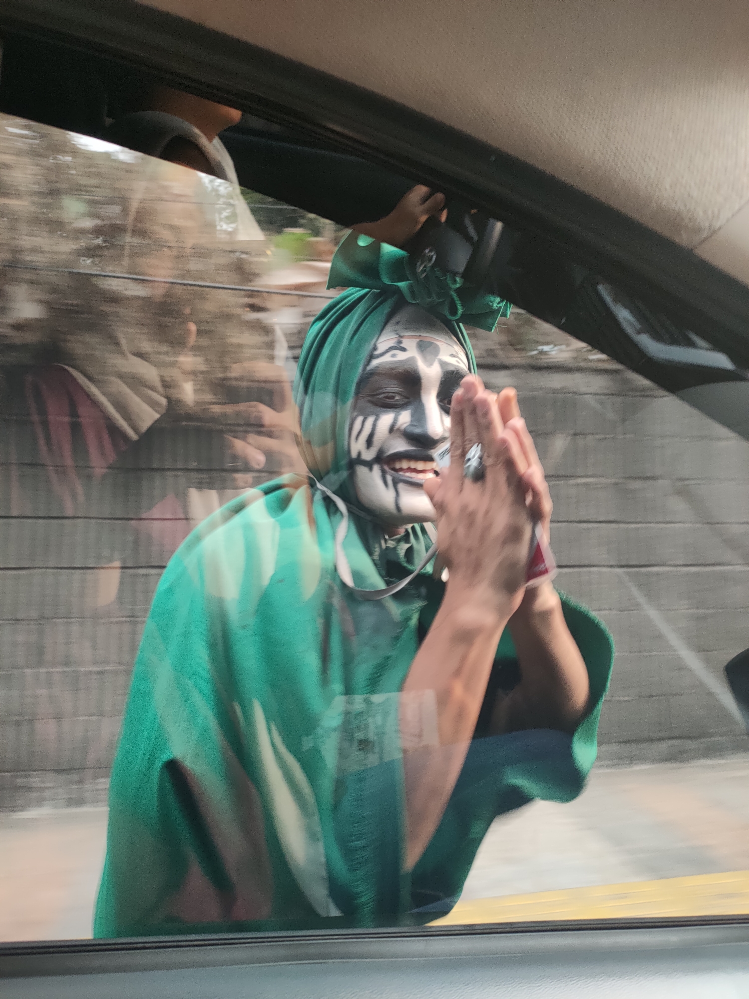 A person with face paint in a green garment is seen from inside a vehicle, looking out a window. The face paint is black and white, resembling tribal markings. The person is wearing a green headscarf and a green cape or robe. Their hands are clasped together in front of them, and they are smiling. Reflections in the window show trees and what appears to be a building with horizontal wooden planks. The image appears to be taken during the daytime, with natural light visible. The setting is outdoors, and the activity seems to be the person interacting with someone or something outside the vehicle, possibly for a performance or demonstration. The location is Lembang, Indonesia.