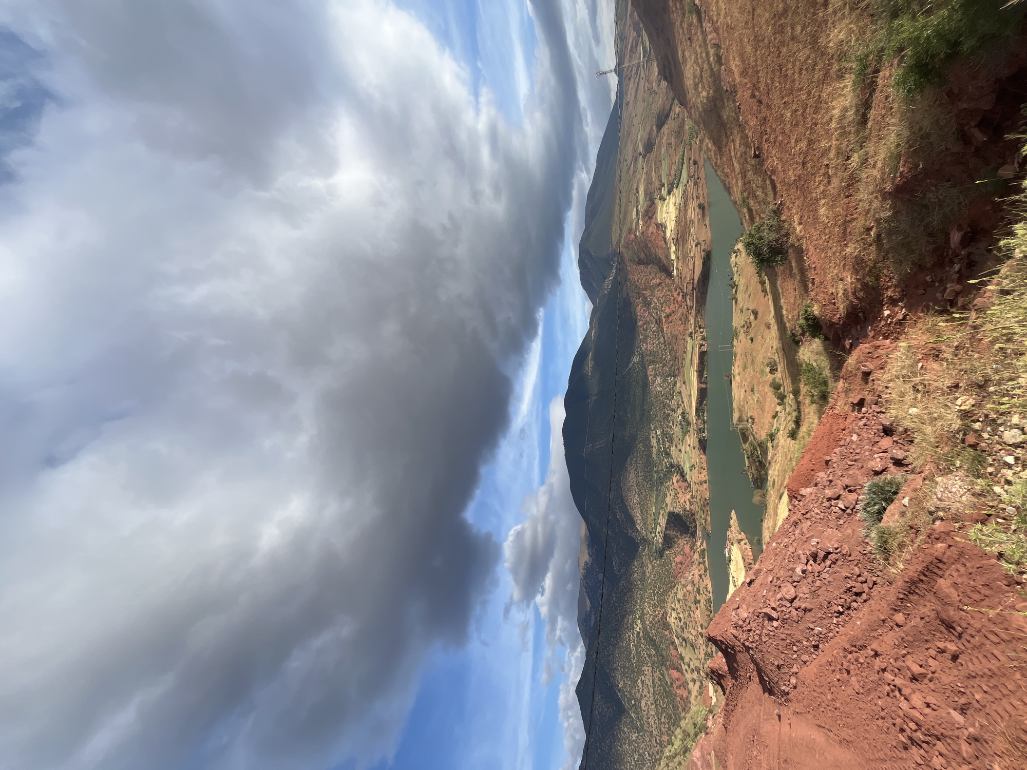 The image depicts a mountainous landscape with a river running through a canyon in the vicinity of Ouaouizert, Morocco. The soil and rock formations exhibit a distinct reddish-brown hue. Sparse vegetation dots the landscape, with some green patches visible. The sky is filled with a mix of dark, grey storm clouds and patches of blue sky. A transmission tower is visible along a distant ridge. A path or road appears in the lower right corner, composed of the same reddish-brown material.