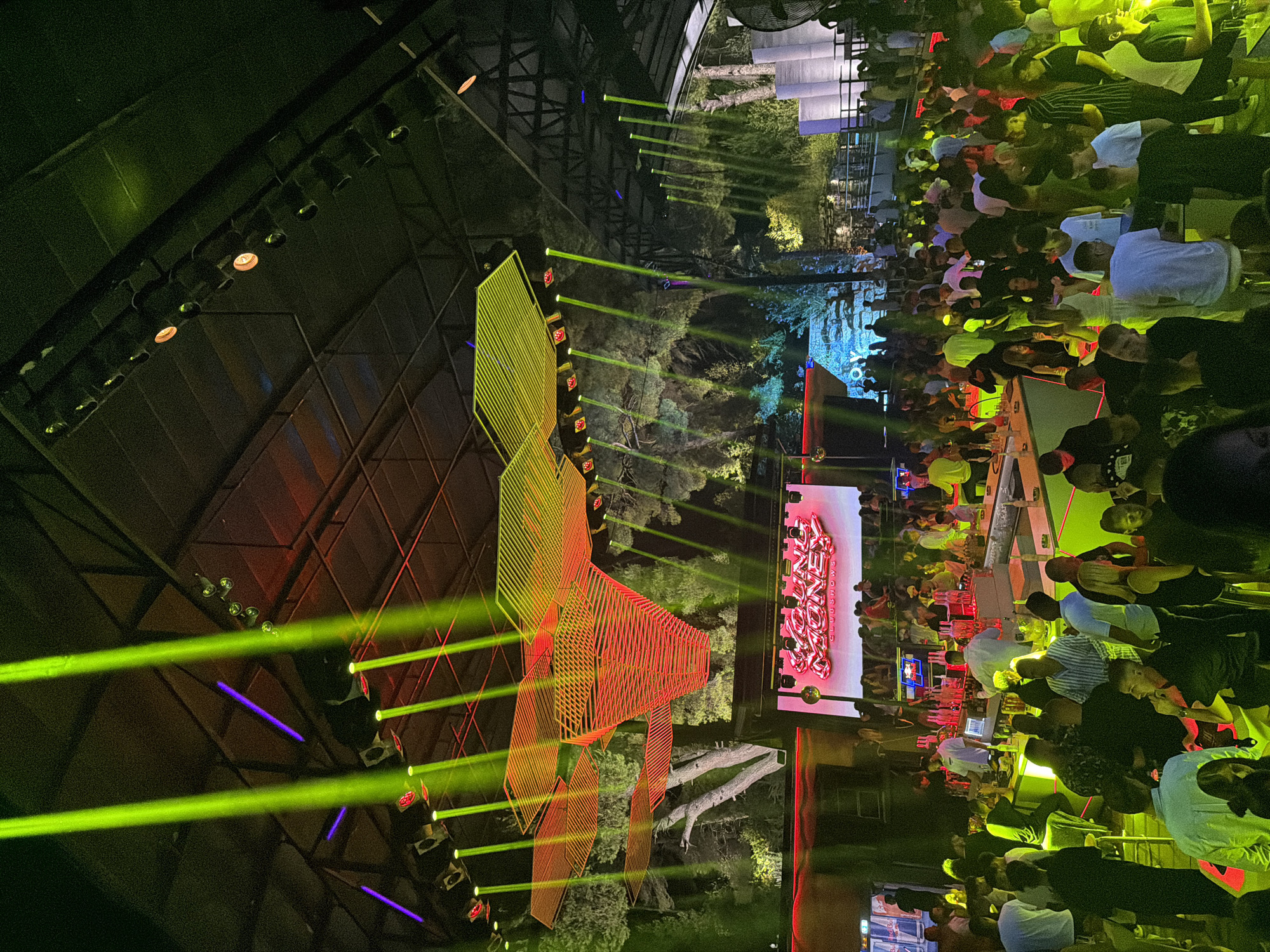 The image depicts a lively indoor party or club scene, likely in Kassandra Municipal Unit, Greece, given the context of the surrounding images. A large crowd of people are gathered, many standing and mingling. There's a prominent bar area with staff serving drinks, indicated by bottles and glasses. Overhead, dynamic lighting effects are visible, including bright green and yellow beams of light cutting through the darkness, illuminating the ceiling structure and a large, multi-layered decorative element. A large screen in the background displays a logo that appears to say "YOUNG MONEY CLUBSHOWS" in a metallic, pink font. The environment suggests a modern entertainment venue with a focus on music and social gathering. Trees are visible behind a screen on the left, suggesting an outdoor or semi-outdoor setup, or a projection that mimics an outdoor setting.