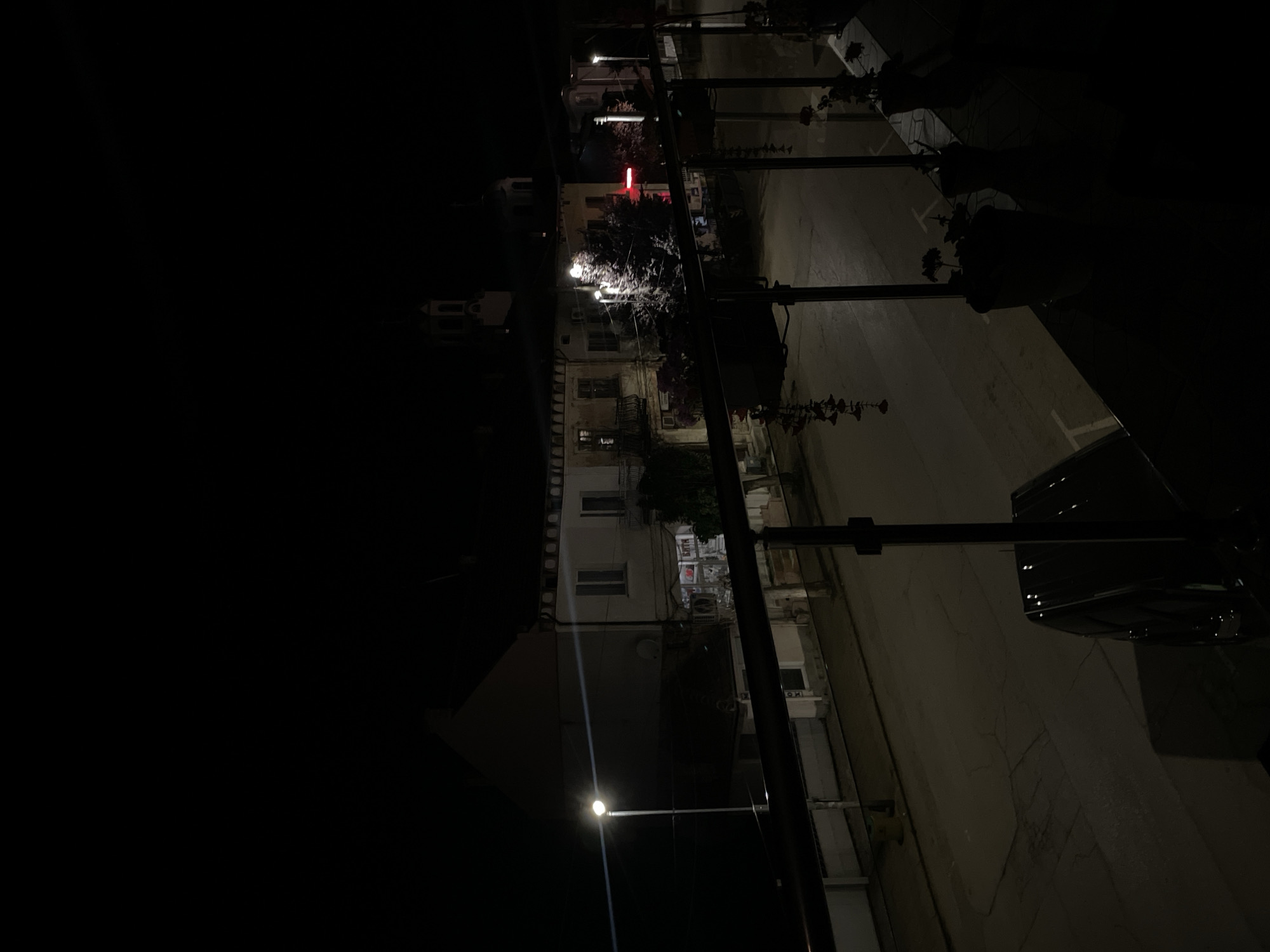 This is a nighttime, eye-level outdoor shot taken from a balcony overlooking a street in the city of Pavlikeni, Bulgaria. A railing with hanging decorative items is in the foreground. Across the street, buildings with illuminated windows and signs are visible, along with streetlights casting pools of light on the pavement. A lone car is parked on the street. The sky is completely dark.