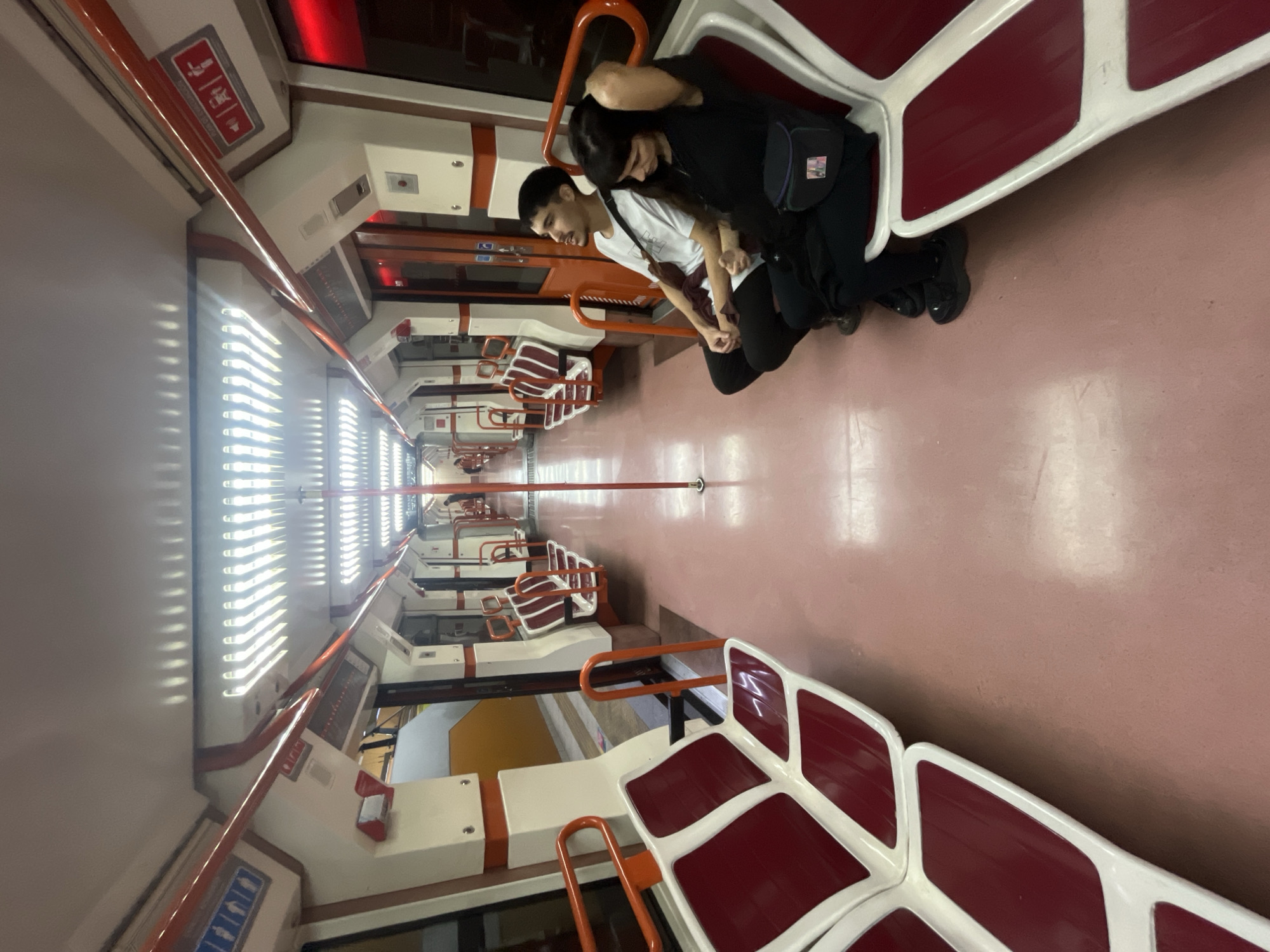 This image shows the interior of a subway car in Buenos Aires, Argentina. Two people are seated together on a bench facing the camera. The person on the right is wearing a black shirt and a small black bag. The person on the left is wearing a white shirt. The subway car is empty except for the two people in the foreground. The walls of the car are white, and the floor is pinkish-red. The seats are a combination of white and burgundy colors. Several rows of empty seats and corresponding orange bars extend into the distance. Overhead fluorescent lighting illuminates the scene.