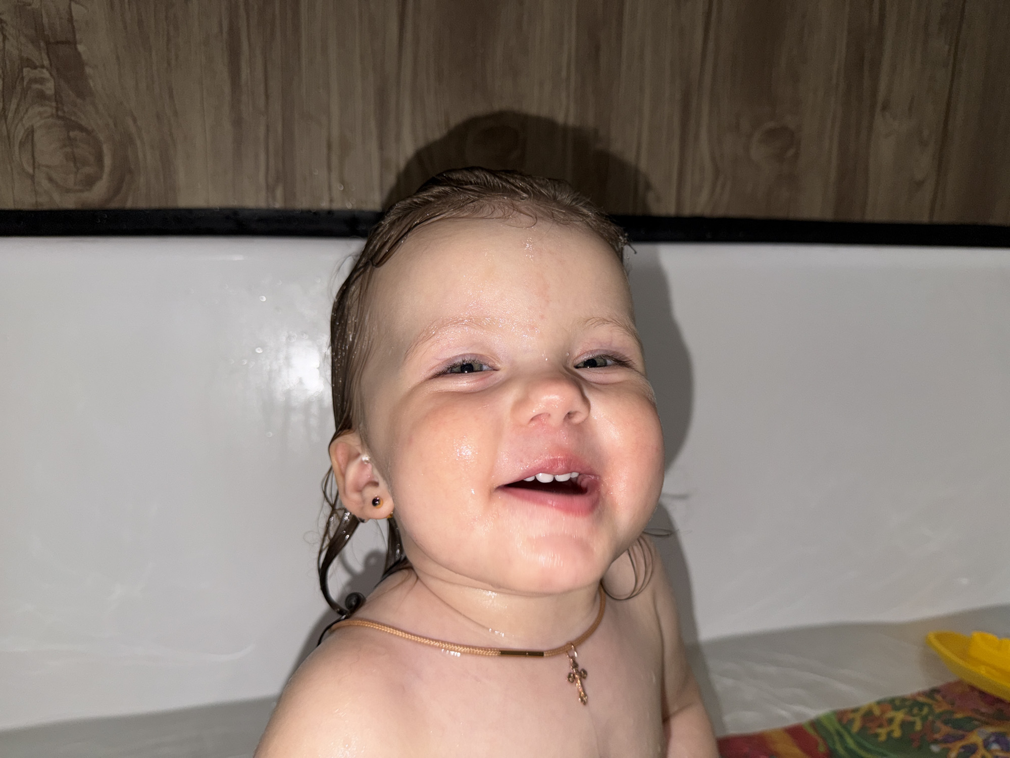 A young child is in a bathtub. The child has wet hair and is smiling. The child is wearing a gold necklace with a cross pendant and earrings. The bathtub is white and has a wooden panel behind it. Part of a yellow toy is visible near the bottom right corner of the frame. There is no indication that this is in Reshetnikovo, Russia.