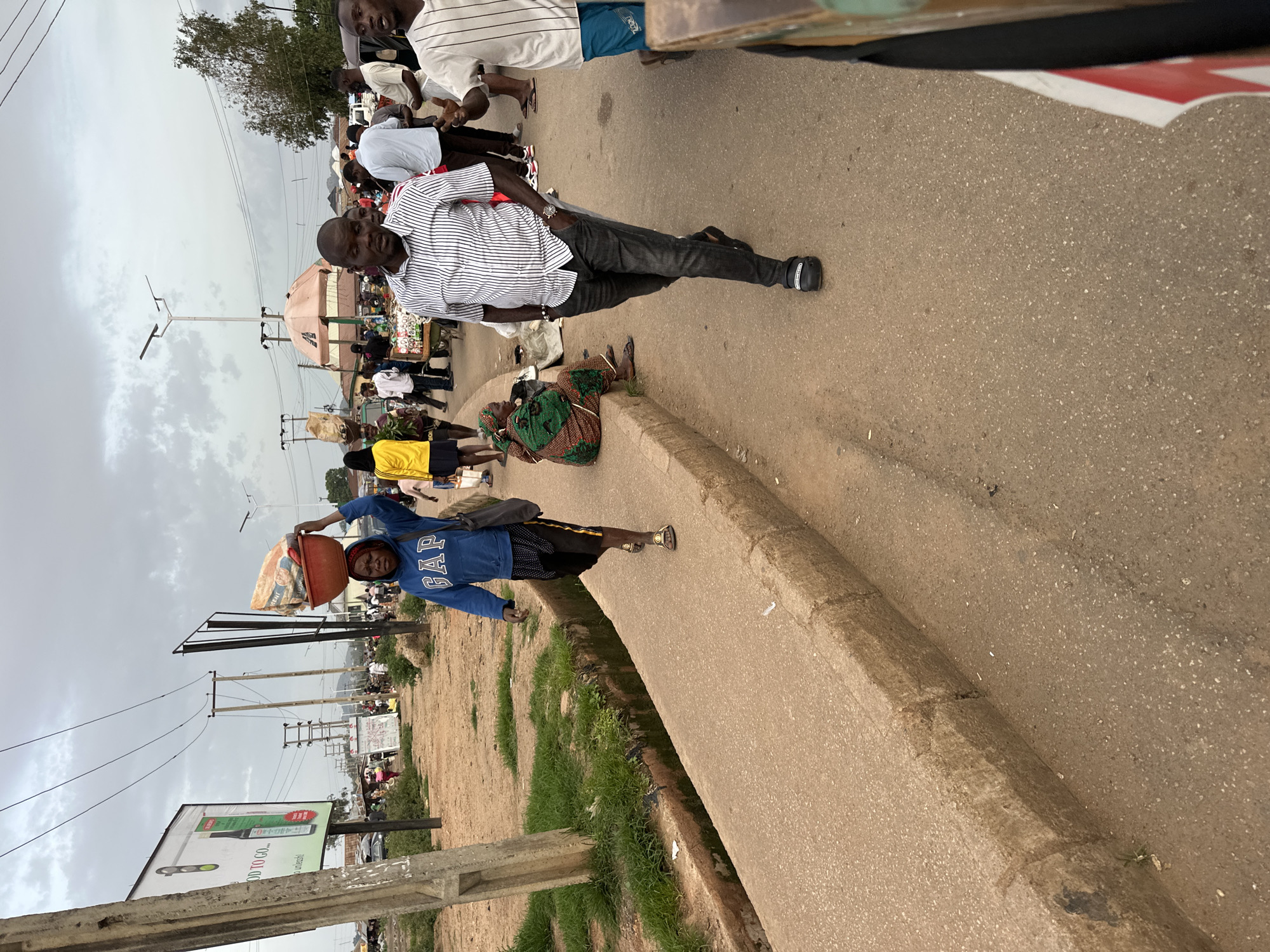 A street scene in Bukuru, Nigeria, shows a woman walking on a sidewalk, carrying a red bowl filled with goods on her head. She wears a blue GAP hoodie and black shorts with white polka dots. A man in a striped shirt and grey pants walks on the road beside her. In the background, other people are visible, some carrying items and others standing near market stalls under an overcast sky. The environment includes a concrete sidewalk, a dirt shoulder, and a road with a drainage ditch. A billboard promoting "GOOD TO GO OIL" is visible on the left.