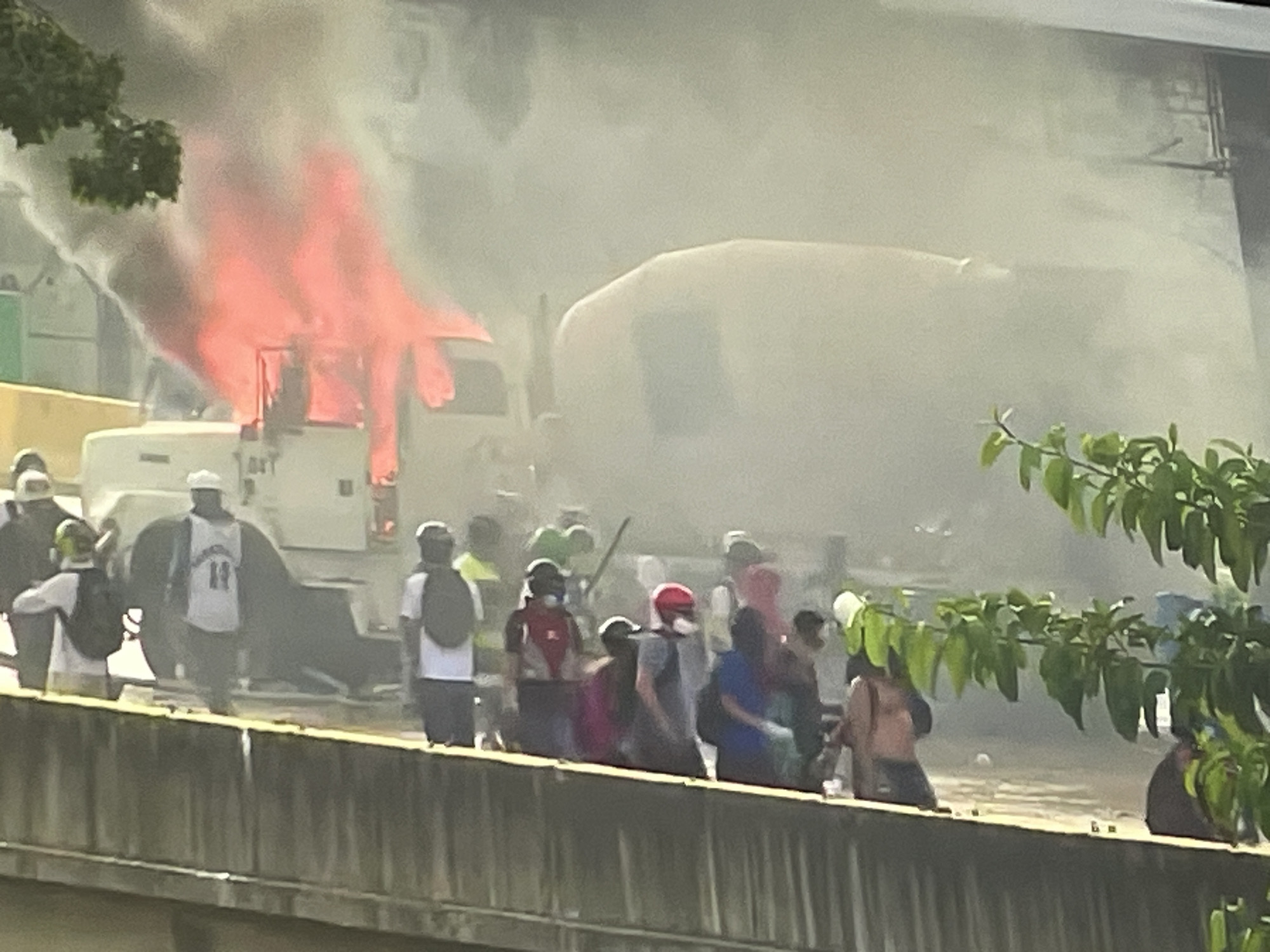 Here is a description of the image:
The image depicts a burning white truck, with flames engulfing the front portion. A large, white cement mixer truck is positioned next to the burning vehicle. A crowd of people stands behind a low concrete barrier, observing the scene. Some individuals are wearing masks. The surrounding environment is obscured by smoke. Green foliage is visible on either side of the image frame. The location is Unknown, Unknown.