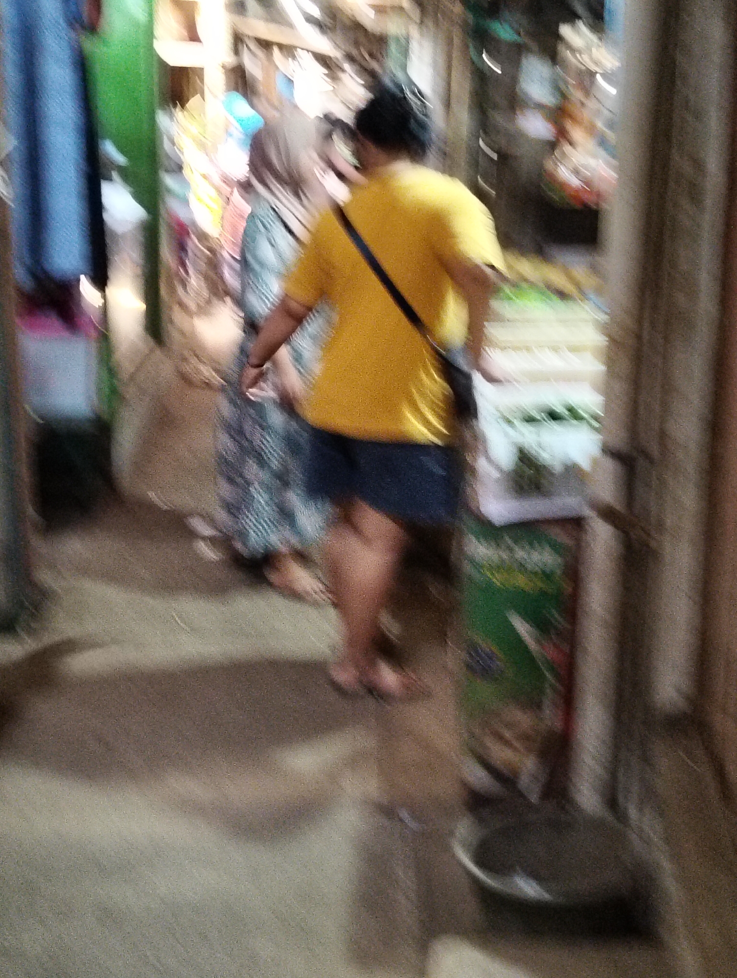 The image depicts a blurred, motion-induced scene within a narrow outdoor marketplace stall area, potentially in Subah, Indonesia, given the context provided. Two individuals are visible, walking away from the camera. The person in the foreground, on the right, is wearing a bright yellow t-shirt and dark shorts, with a dark strap across their chest. The person to their left is wearing a patterned dress. The environment is characterized by narrow walkways and the presence of various market stalls, indicated by blurred structures and items for sale in the background. The lighting is somewhat dim, contributing to the overall blurriness and the indistinct nature of the details within the market. The ground appears to be a sandy or dirt path.