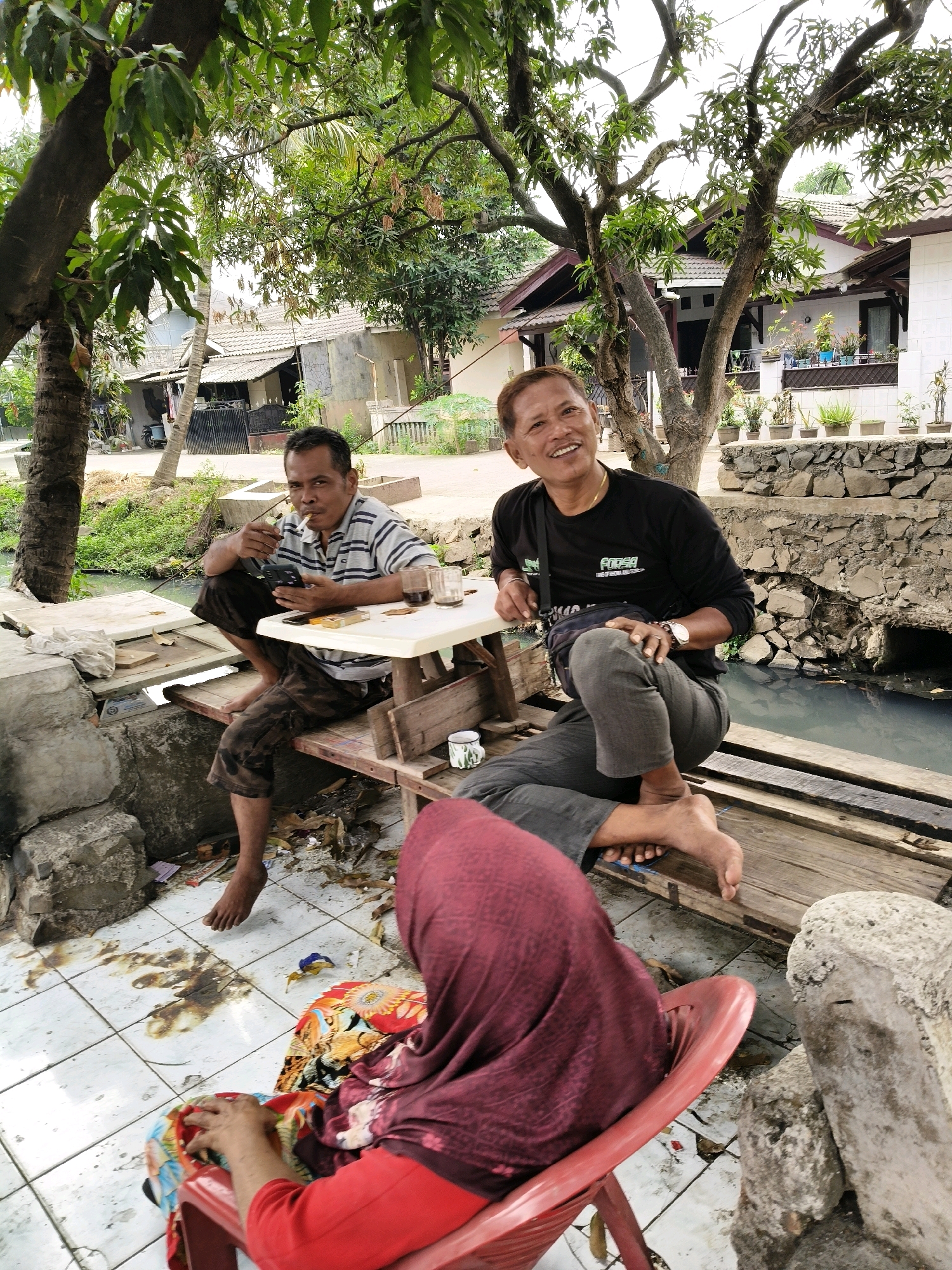 In Setiamekar, Indonesia, three individuals are present in an outdoor setting near a body of water. One man, wearing a striped shirt and camouflage pants, is seated on a wooden bench to the left, smoking a cigarette and holding a mobile phone. He has a pack of cigarettes and a lighter on the table. To his right, another man in a black long-sleeved shirt is seated on a similar wooden bench, smiling and looking upwards. He has a bag slung over his shoulder and is wearing a watch. A third individual, whose back is to the camera and is wearing a red shirt and a maroon headscarf, is seated in a red plastic chair in the foreground, facing away from the other two men. The environment includes tiled flooring, a small table between the men, a stone wall, vegetation, and houses in the background. There appears to be debris on the ground.