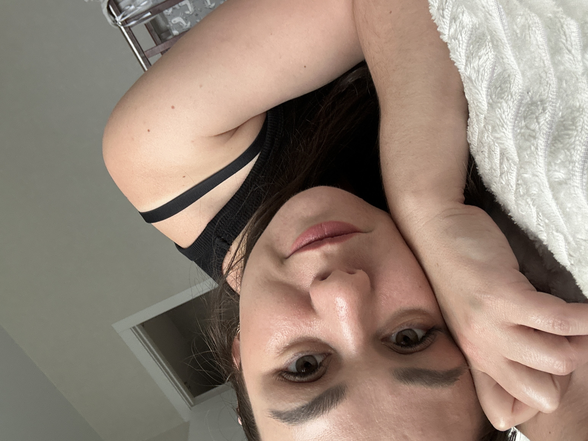 A woman is lying down, her head resting on her hand and a white, textured object, possibly a towel. She is wearing a black tank top with thin straps. The location is unclear, as the image does not provide enough information to confirm the specific location in Chaikovsky, Russia, or any location at all. The visible background features a white ceiling with an access hatch and a metal rack.