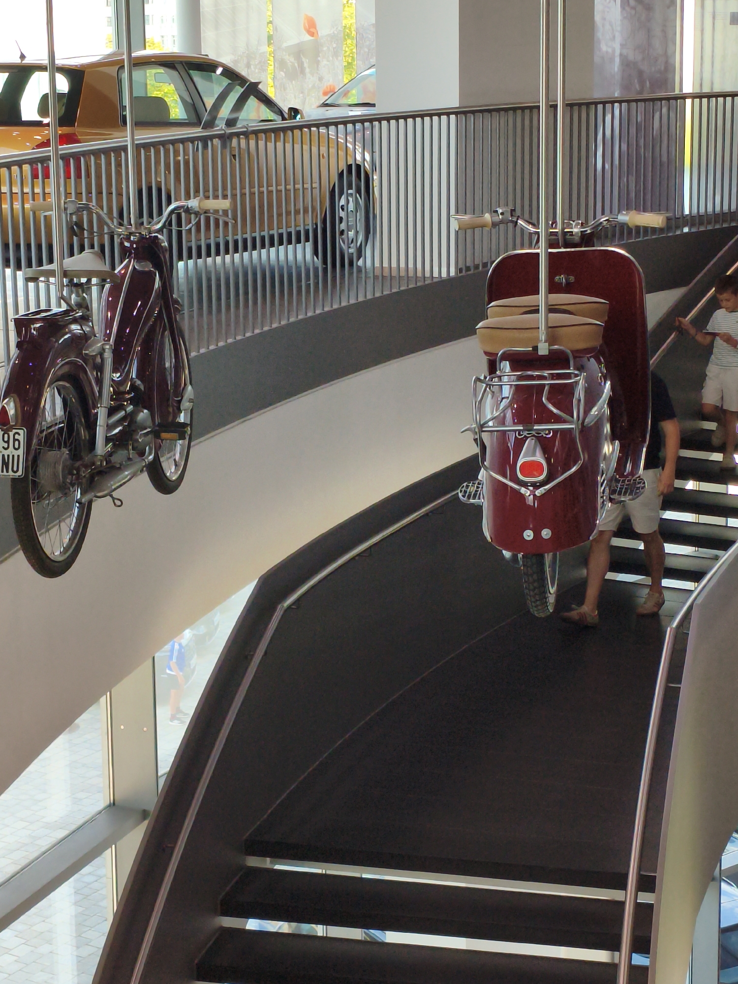 The image shows an interior view of the Audi Forum in Ingolstadt, Germany, which includes a museum exhibition. In the foreground, a maroon vintage scooter is suspended mid-air by metal poles. The scooter has a chrome luggage rack on the front and two beige seats. Further down the suspended display, another similar maroon scooter is also hanging. In the background, a section of a dark-colored staircase with metal railings is visible, leading upwards. On the staircase, a young boy wearing a striped t-shirt and white shorts is walking upwards. To the left of the staircase and scooters, a golden vintage car is parked behind a metal railing. The environment appears to be a modern building with large windows, suggesting a showroom or museum space. The overall scene depicts a display of vintage vehicles within a contemporary architectural setting.