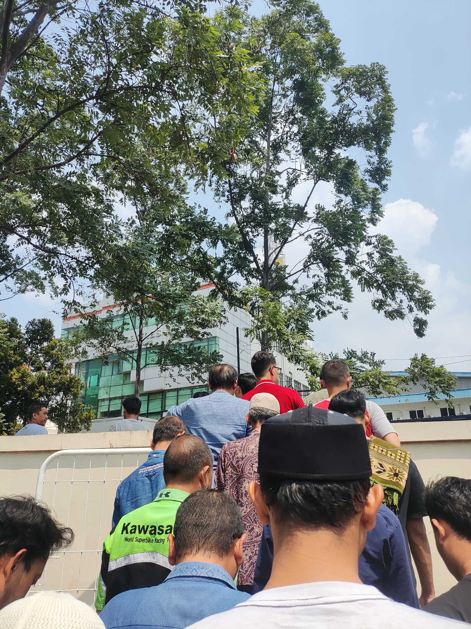 A group of men are gathered outdoors, standing in a line and facing away from the camera. They appear to be waiting or observing something. In the background, a modern, multi-story building with green-tinted windows is visible, surrounded by lush green trees. The sky is blue with scattered white clouds. The scene is likely a public space in East Jakarta, Indonesia, possibly near an office building or an event.