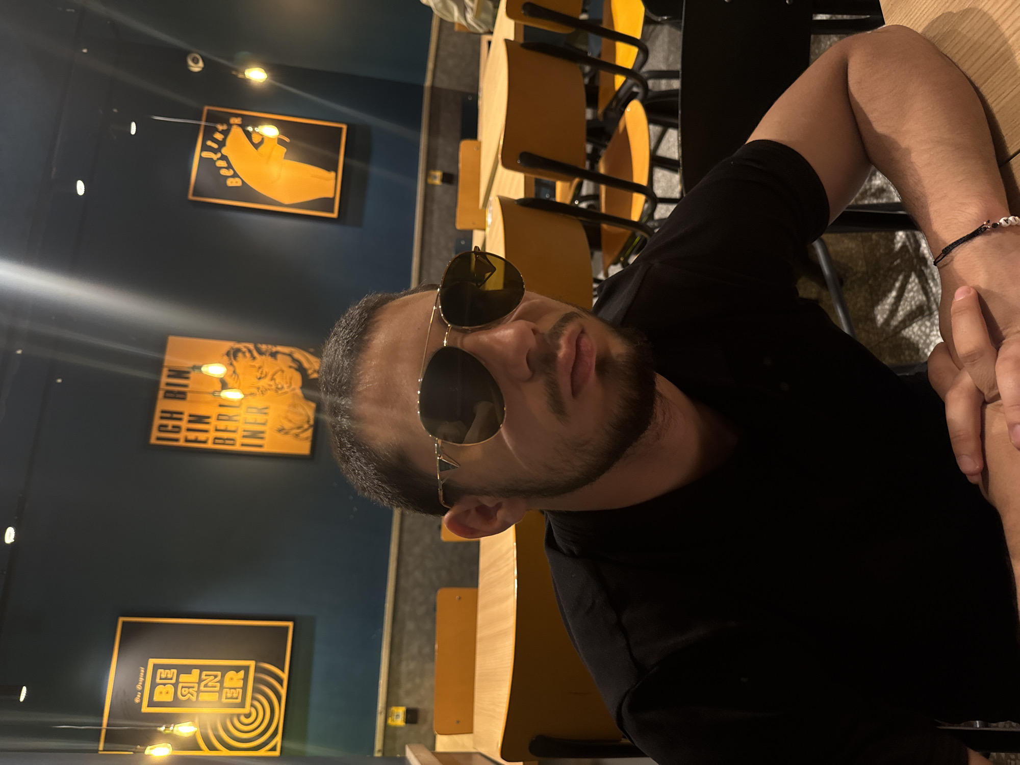 A medium shot of a man with a beard, wearing sunglasses and a black t-shirt, sitting at a table in a restaurant or café. The man is fair-skinned. There are other chairs visible. The walls of the establishment are blue, and there are framed pictures on the wall including a picture of an hand. It is believed the image was taken in Strasbourg, France, as the images are related to Berlin.