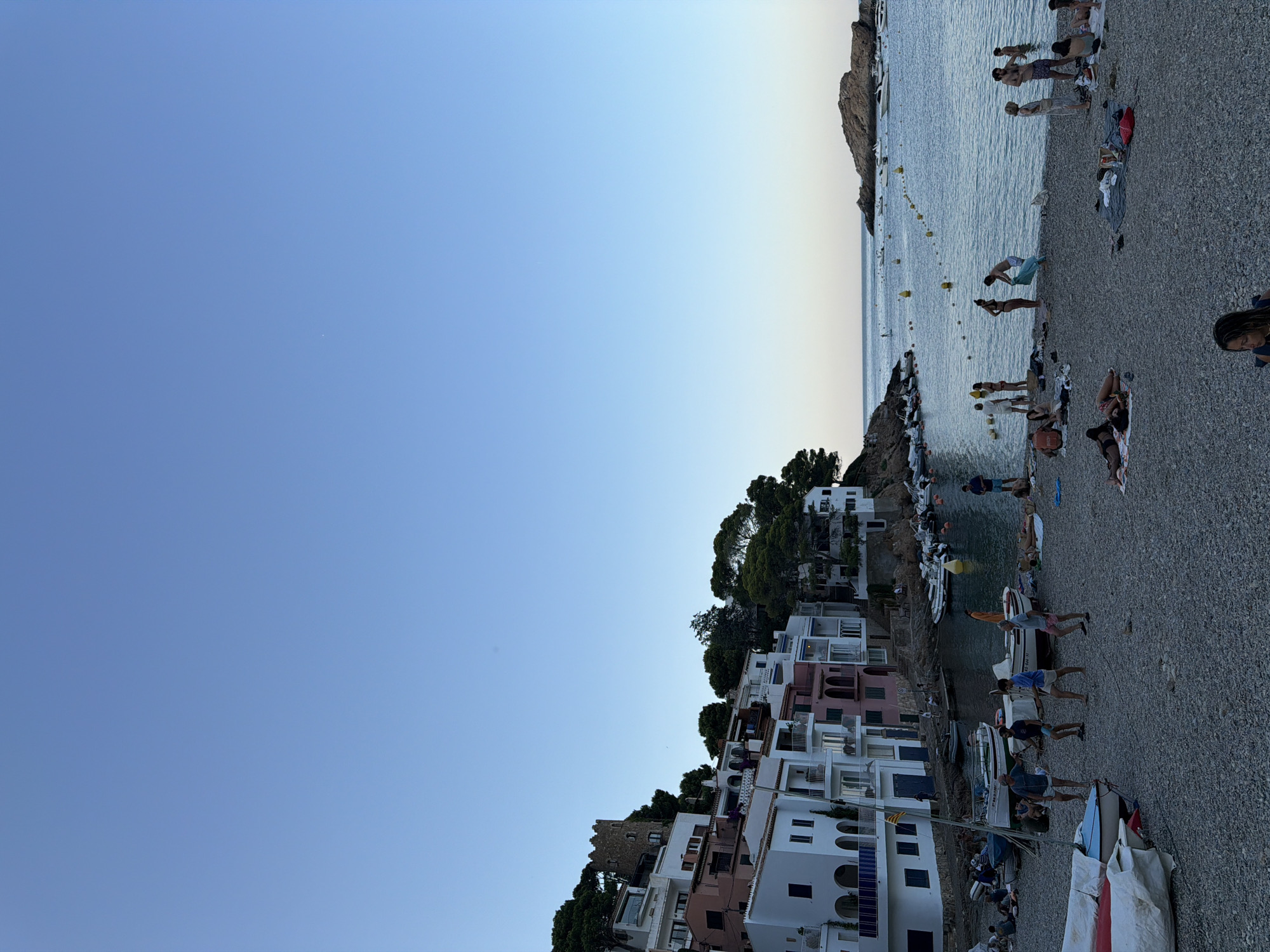 The image depicts a coastal town in Begur, Spain, characterized by its picturesque Mediterranean architecture. White and pastel-colored buildings, adorned with balconies and arches, cascade down a hillside towards a pebble beach and a calm, blue bay. Several small boats are moored in the water, and a few larger yachts are visible in the distance. The beach is populated with people, some sunbathing on towels, others wading in the shallow water, and a small group engaged in a casual game on the shore. Lush green trees dot the landscape, providing a natural contrast to the built environment. The sky is a clear, pale blue, indicating a pleasant, possibly late afternoon or early evening atmosphere. In the distance, a rocky island rises from the sea, adding to the scenic beauty of the location. A medieval tower stands prominently on the hillside, hinting at the historical significance of the area.