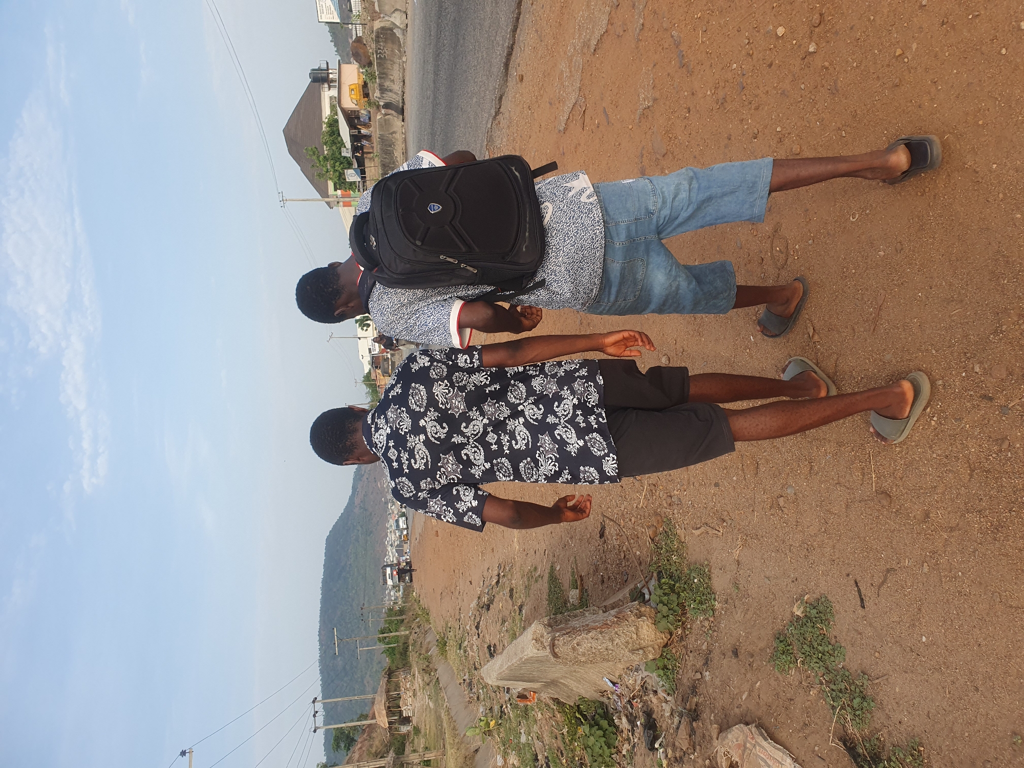 Two young men stand on a dirt and gravel shoulder beside a road. One wears a light blue shirt with a pattern, denim shorts, sandals, and a black backpack. The other wears a dark shirt with a white pattern, dark shorts, and sandals. The road is paved, and a building with a slanted roof and some shops are visible in the background. In the distance, there is a mountain. The location is in Lokoja, Nigeria. There is trash, concrete debris, and greenery on the shoulder.