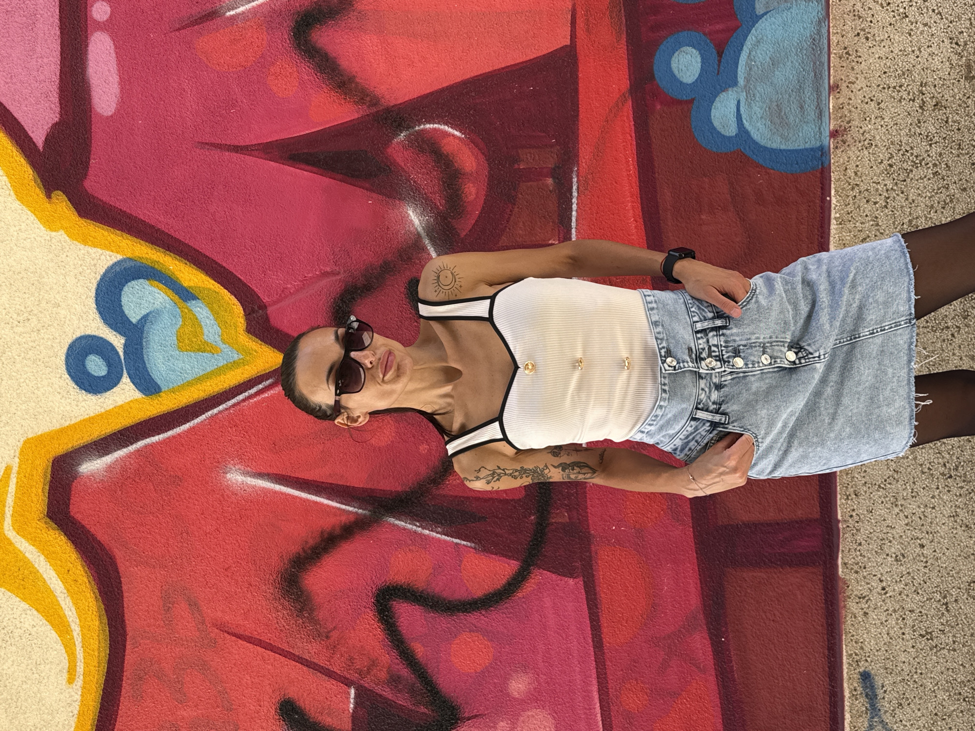 A young woman is captured in a full-body portrait, posing against a vibrant, graffiti-covered wall. The image is rotated 90 degrees clockwise, making her appear to be lying down. She looks upwards towards the camera, wearing dark sunglasses that obscure her eyes, and has a composed, neutral expression with a hint of a smile.
She is dressed in a white ribbed tank top with black trim and gold-colored buttons down the front, paired with a light-wash denim mini skirt featuring a button-up closure and a frayed hem. She also wears sheer black tights or stockings. Accessories include a dark wristwatch on her left wrist, a delicate chain necklace with a small pendant, and a bracelet on her right wrist. Visible tattoos include a stylized sun and moon design on her right upper arm and a detailed floral or vine-like pattern on her left forearm. Her dark hair is pulled back.
The background is a vividly painted wall dominated by abstract graffiti in shades of red, pink, yellow, blue, and black, suggesting an urban outdoor setting in Pleven, Bulgaria. A light-colored concrete or paved surface is visible to the right, indicating the ground. The scene is brightly lit, indicative of a sunny day. No legible text is visible in the graffiti.