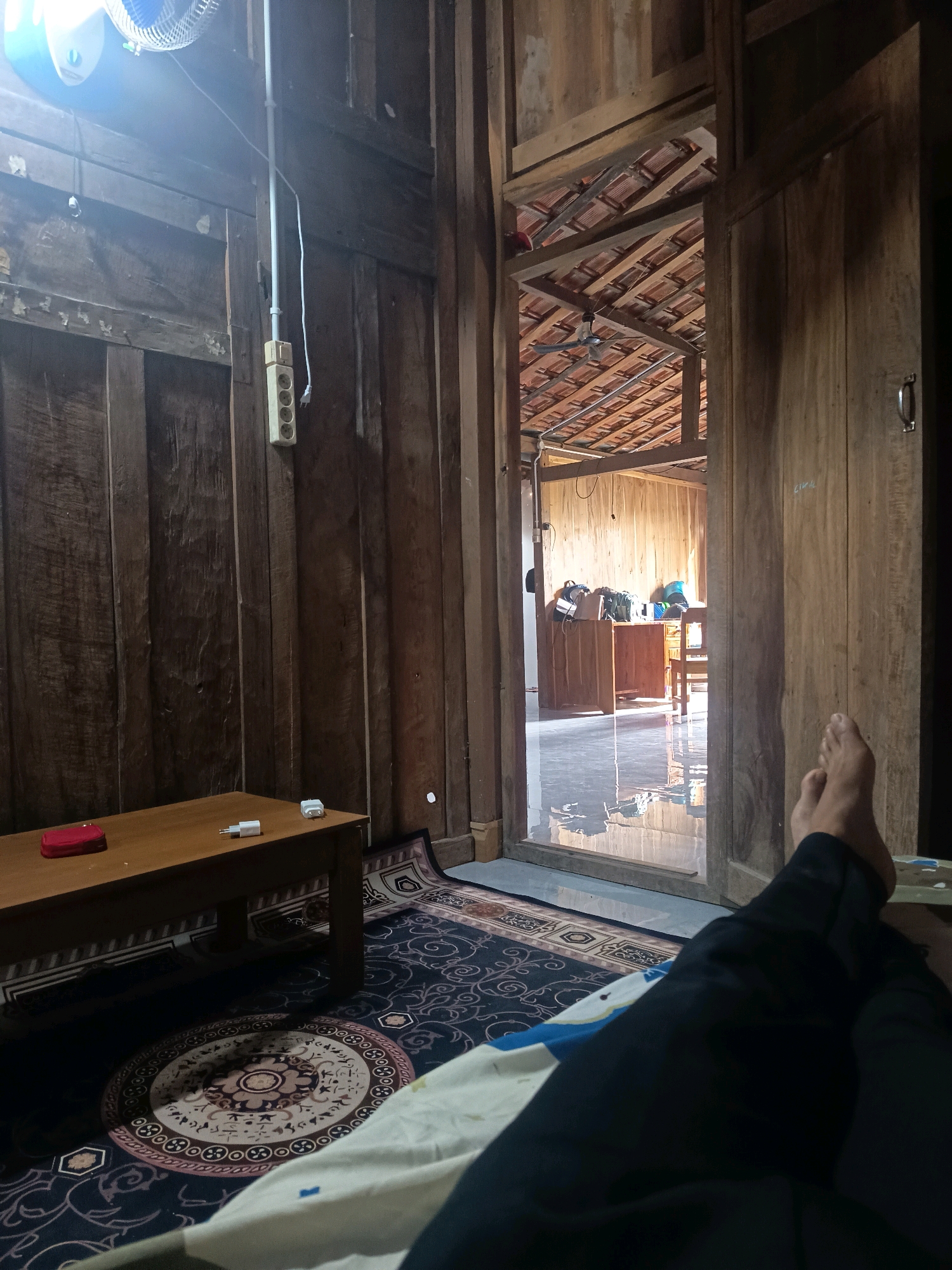 The image depicts the interior of a room constructed with dark wood paneling, characteristic of some traditional Indonesian homes, potentially located in or near Sidorejo, Indonesia. The room features a dark-colored carpet with intricate circular and geometric patterns on the floor. A wooden table is positioned on the left side of the room, with a red rectangular object and two white electrical adapters on its surface. A ceiling fan is visible in the upper left corner of the image, emitting a bright light.
An open doorway on the right side of the frame reveals another room with a polished floor that reflects light. This adjacent space contains a wooden desk and chair, with several bags placed on the desk. The ceiling of this area is visible, made of reddish-brown tiles supported by wooden beams.
In the foreground, a person's legs are visible, clad in dark pants, with their feet crossed. The perspective suggests the person is lying down or relaxing, possibly on a bed or a hammock that is mostly out of frame. The overall impression is one of a simple, possibly rural, living space.