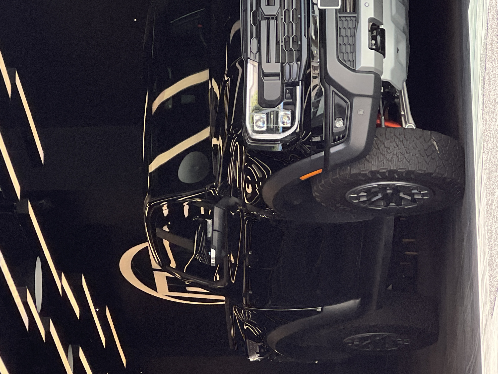 A flipped image shows a black vehicle, possibly a truck or SUV, parked indoors, likely a showroom or display area. The front of the vehicle, including the grille, headlights, and front tire, is prominently visible. The vehicle is parked on a black surface bearing a logo, possibly the vehicle manufacturer's or a dealership's. The background features vertical light patterns, likely from spotlights or architectural lighting. Based on the image information, the location in the city of Tanger طنجة, Morocco, cannot be determined.