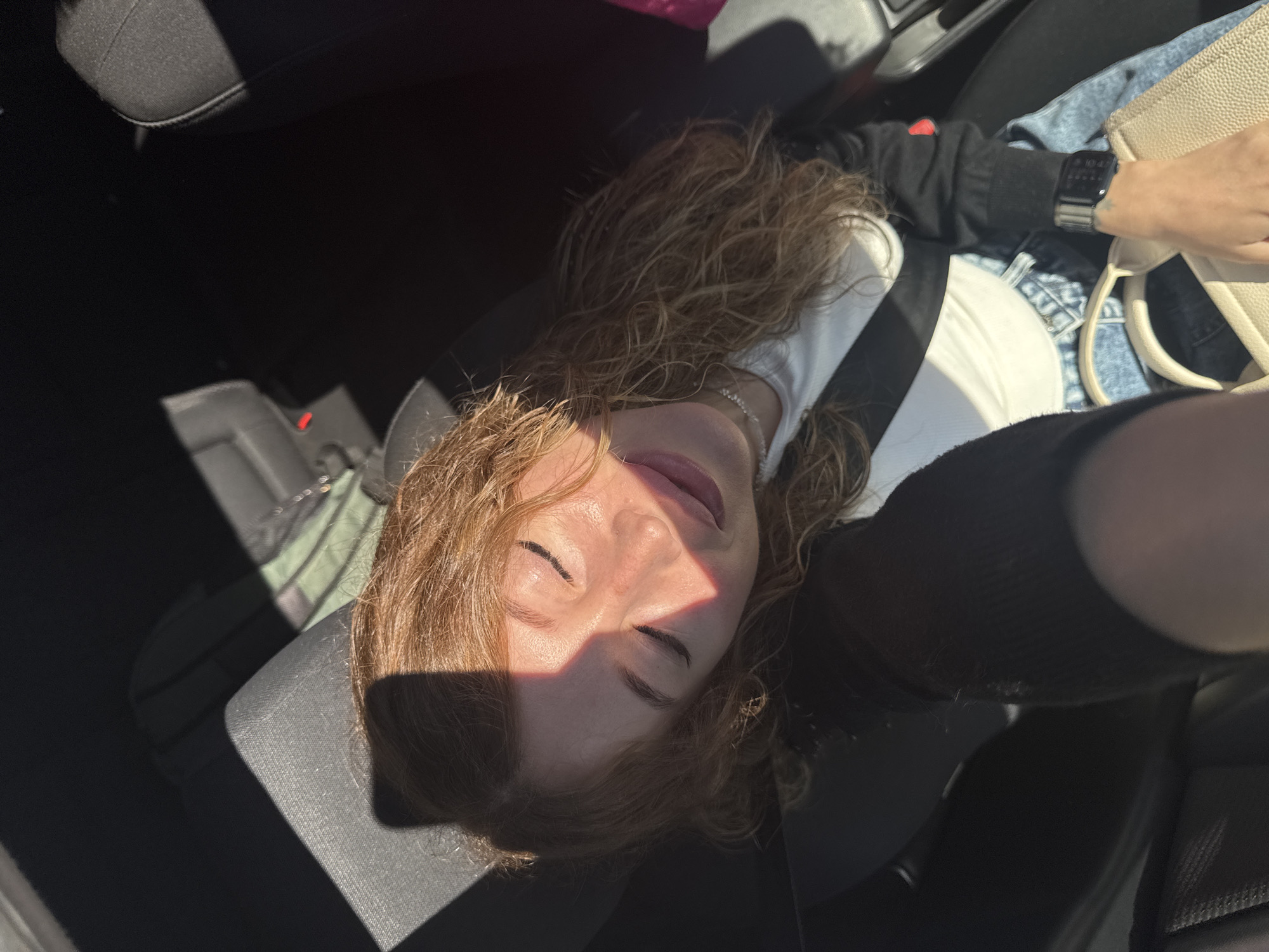 A high-angle, medium shot captures a young woman with auburn, wavy hair sitting in the passenger seat of a car. Sunlight casts a diagonal shadow across her face, highlighting her closed eyes and a soft smile. She wears a white top, a black zip-up jacket, and blue jeans. A seatbelt is fastened across her chest. A light-colored handbag rests on her lap, and a smartwatch is visible on her left wrist. The interior of the car appears to be dark with hints of dark grey upholstery. There is no information in the image to suggest a location in Chaikovsky, Russia.