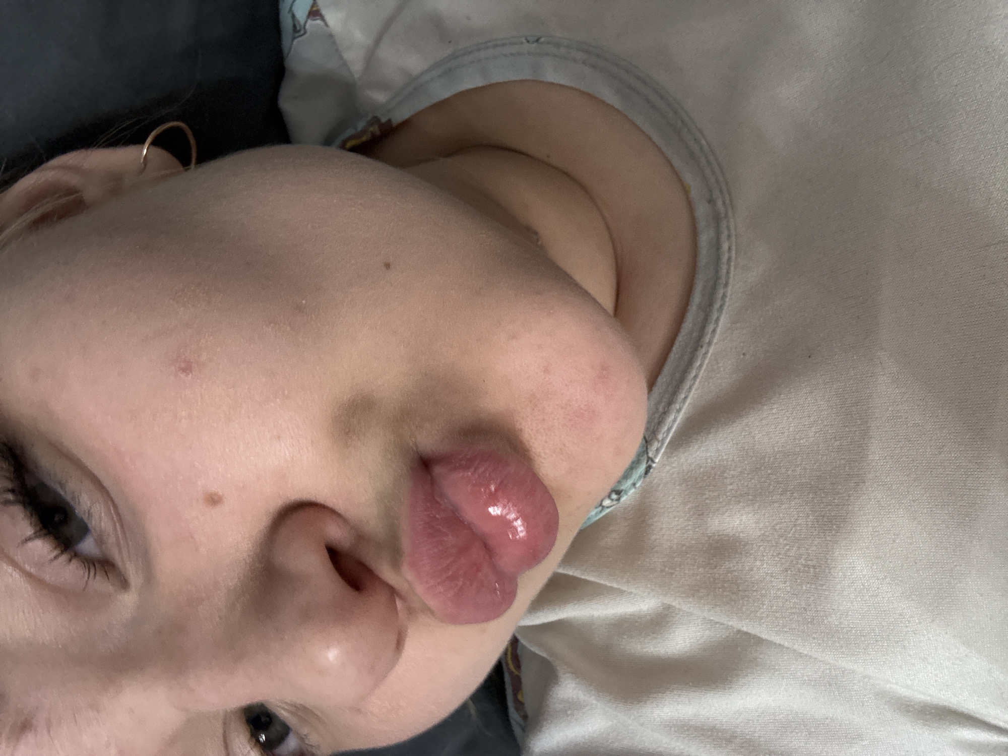 A close-up, rotated view shows a person's face, likely a child, with puckered lips. The person has fair skin, brown eyes, and dark eyelashes. A gold hoop earring is visible in the ear. They are wearing a light-colored shirt or top with a light blue trim. The background consists of a dark gray surface. The location is unknown.