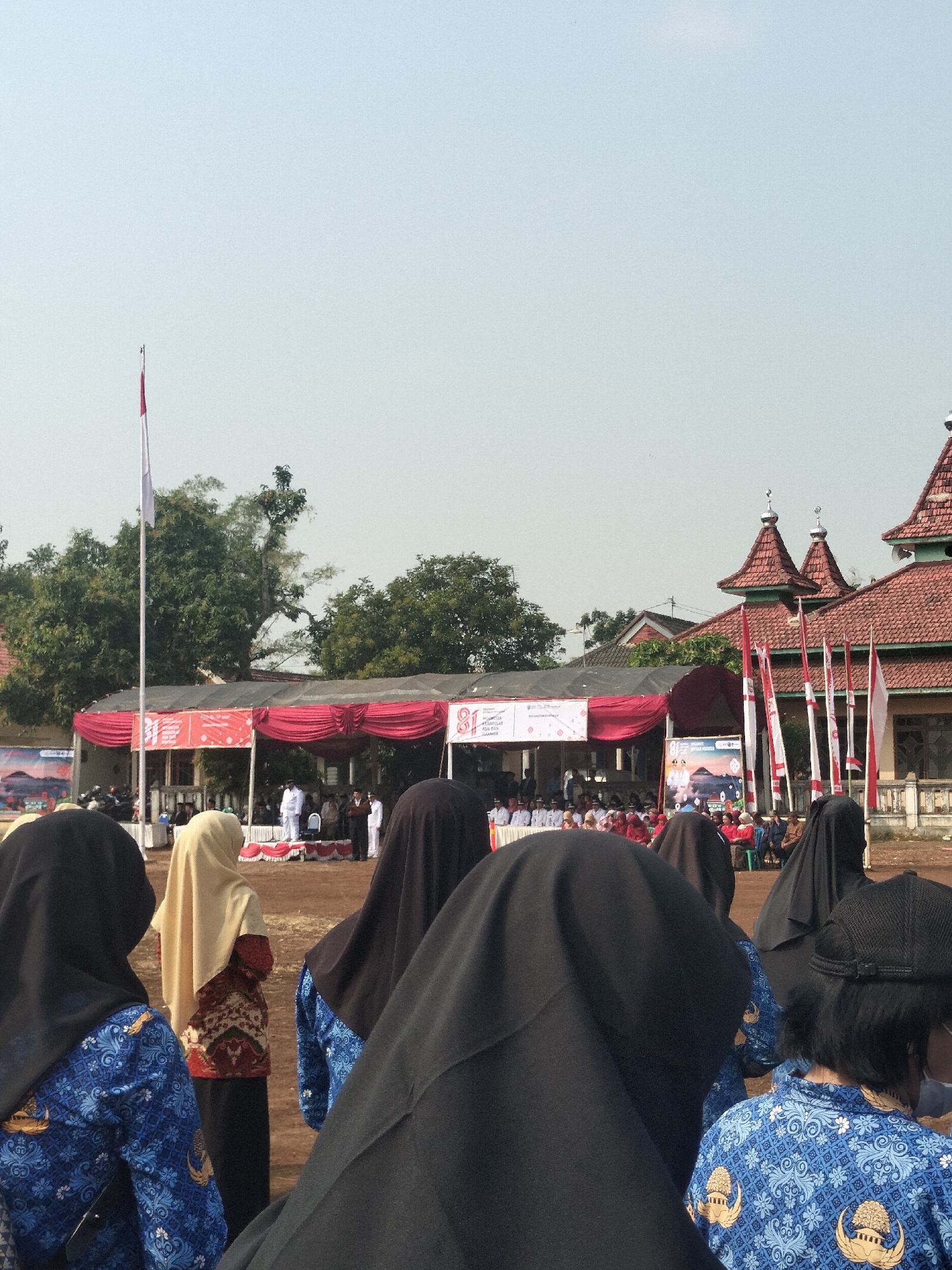 In Bantaran, Indonesia, this image captures a public gathering. In the foreground, several individuals, predominantly women wearing headscarves, are seen from behind. Some are dressed in blue uniforms with intricate white patterns. One woman in the center wears a tan headscarf and a patterned top. In the mid-ground, a stage is set up with red drapery and banners displaying text and imagery, including the number "81". Several people are visible on and around the stage, including a man in a white suit. A flagpole with a red and white flag stands to the left of the stage, and multiple Indonesian flags are positioned to the right. In the background, traditional Indonesian-style buildings with red tiled roofs are visible amongst lush green trees. The scene appears to be an outdoor event under a clear, bright sky, likely a ceremony or celebration.