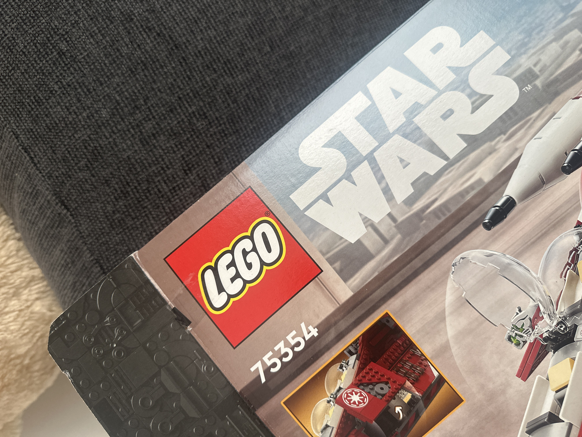 This close-up, angled image showcases a LEGO Star Wars set box, resting against a dark, textured fabric surface, likely a sofa or cushion, in an indoor setting in Sofia, Bulgaria.
**Subjects:** The primary subject is a LEGO box. The dark fabric object serves as a background element.
**Objects:** The LEGO box displays the iconic red and yellow LEGO logo, followed by the white text "STAR WARS" with a trademark symbol. Below the LEGO logo, the set number "75354" is clearly visible in white. Portions of the box artwork are visible, depicting a red and white spaceship or vehicle, including a clear cockpit canopy and detailed red brick components of its interior. The left edge of the box features a textured dark grey design.
**Scene/Setting:** The scene is an indoor environment, typical of a home, where the LEGO box is placed on or next to a piece of furniture.
**Activity or event taking place:** No explicit activity is depicted. The presence of the boxed set suggests an impending build, a gift, or storage of the item.
**Notable details:** The image is a static shot with no visible movement. The lighting is soft and even, suggesting either diffused natural light or typical indoor artificial lighting. The fabric background has a woven texture and is slightly out of focus, drawing attention to the sharply detailed box. No specific time of day, weather, or emotions are discernible.
**Visible text:** "LEGO", "STAR WARS", and the set number "75354".