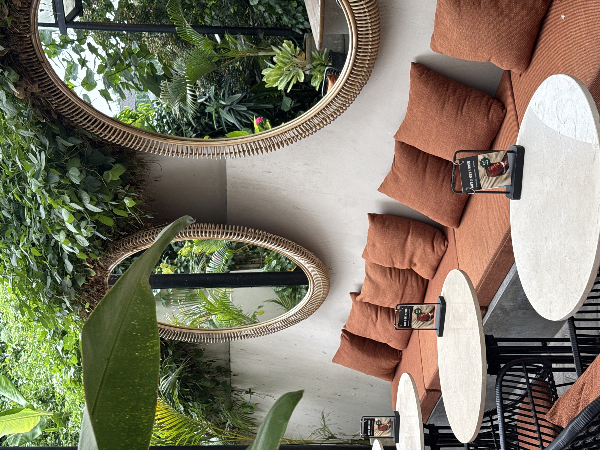 The image depicts an interior space in the Ubud District, Indonesia, featuring a seating area composed of an orange sectional couch and several small, round, light-colored tables. Black wicker chairs surround the tables. Each table has a small sign or advertisement stand on it. Two large, oval mirrors with rattan frames are mounted on the wall, reflecting lush greenery outside. The surrounding environment includes an abundance of plants, adding to the natural and possibly tropical ambiance. There are no people visible in the image. The overall impression is that of a stylish, nature-integrated space, likely a restaurant or cafe.