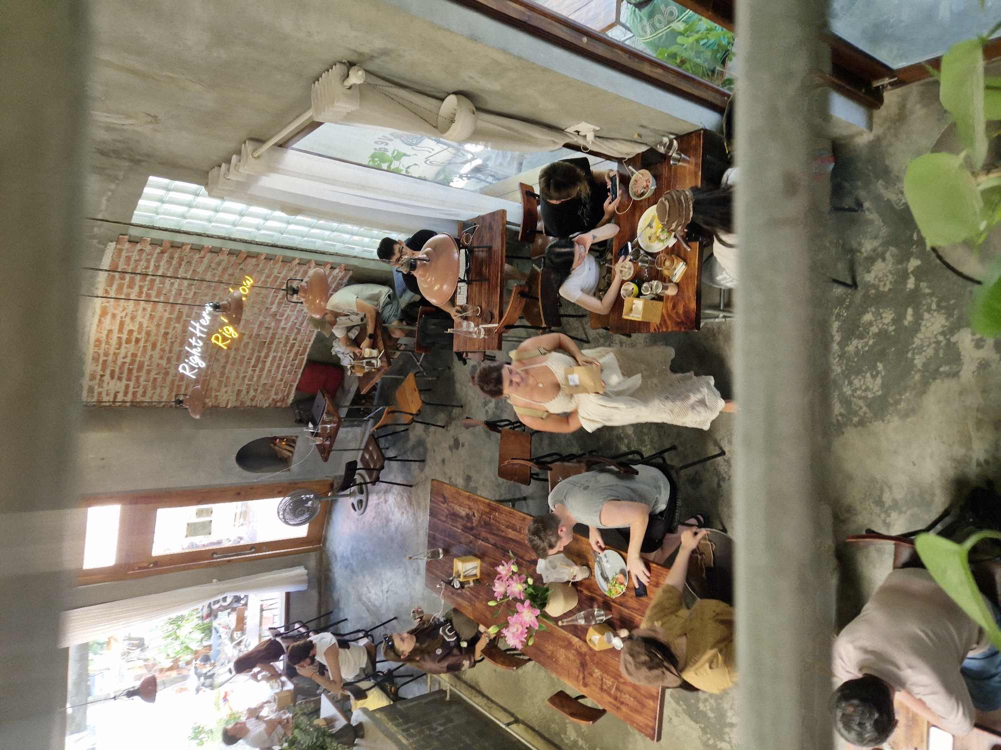 The image depicts an indoor scene within a bustling cafe located in Đà Nẵng, Vietnam. The cafe has a modern, industrial aesthetic, with exposed brick walls, concrete floors, and large windows. Multiple groups of people are seated at various tables, engaged in activities such as dining, conversing, and using their mobile devices.
In the foreground, a group of four individuals are seated around a wooden table. One person, wearing a brown hat and yellow shirt, is reaching across the table. Another person, wearing a grey t-shirt, is looking down at their plate. A woman in a white dress is standing and holding a bag.
Further back, several other tables are occupied. A couple sits at a table near the window, looking at their phones. Another group is gathered around a larger table, with food and drinks spread out. A person is visible at a table in the far background, facing away from the camera.
The cafe is furnished with wooden tables and chairs, as well as some industrial-style lighting fixtures. Plants are strategically placed throughout the space, adding a touch of greenery. The overall atmosphere appears lively and relaxed.