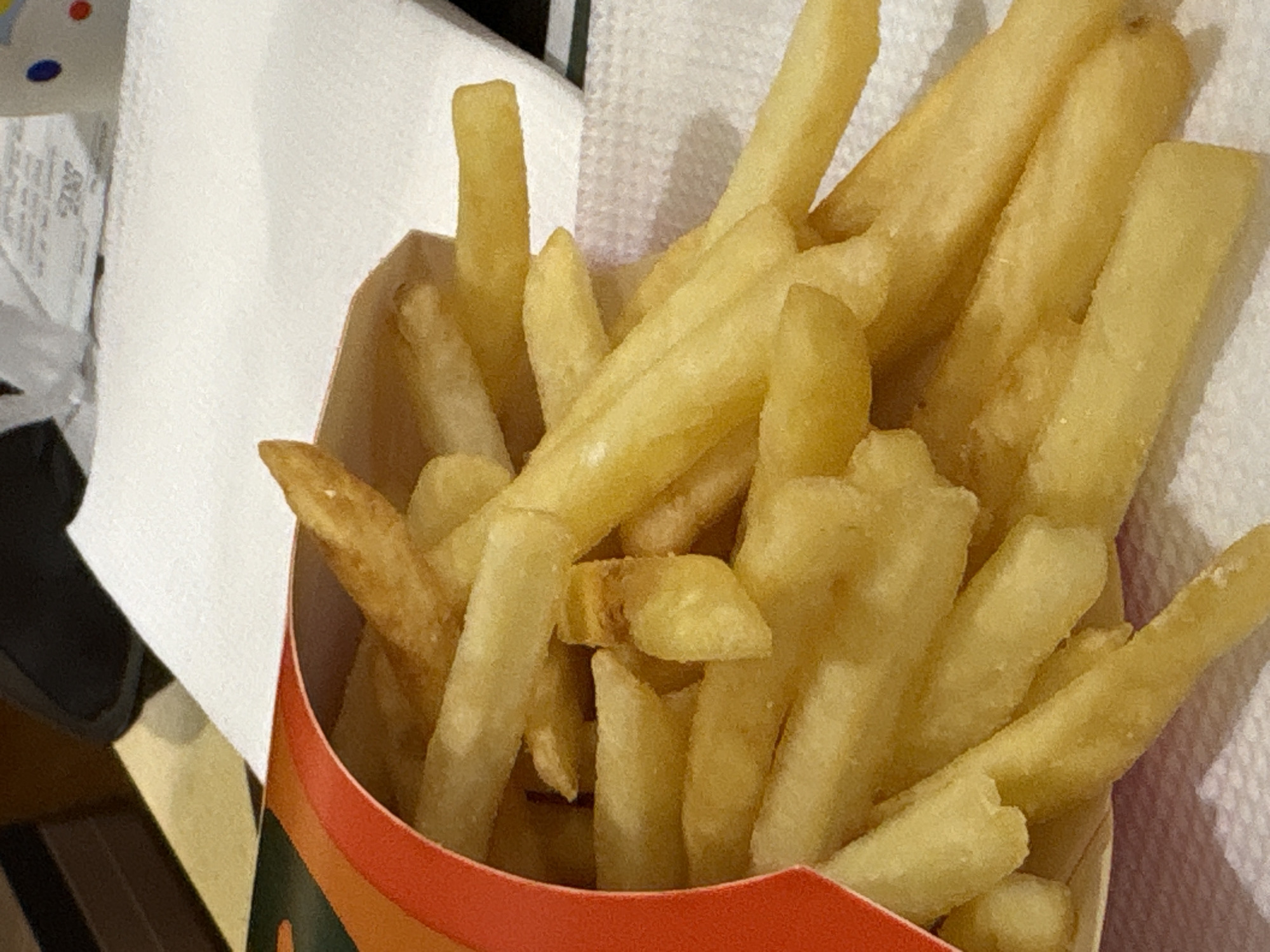 The image presents a close-up view of a container filled with french fries, positioned against a backdrop of white paper material. The primary subjects are the numerous golden-yellow french fries, which are standard-cut and appear lightly cooked, with some exhibiting slight browning. They are piled high within a paper container that features a vibrant design at its upper rim, consisting of horizontal stripes in shades of red and orange, transitioning to green towards the bottom left.
In the background, to the left and extending upwards, a crinkled white paper item is visible, possibly a napkin or a receipt. There is blurry, illegible text printed on this paper, but no specific words or characters can be deciphered. A textured white surface, resembling a paper napkin or towel, lies behind the fries and their container, providing a soft-focus background. Dark, out-of-focus objects are partially visible in the bottom left and top left corners, likely indicating parts of a table, bag, or another surrounding item.
The scene depicts a still life of fast food, suggesting an indoor setting such as a restaurant, a takeout meal at home, or a casual dining environment. No human activity is explicitly shown, but the arrangement implies the fries are either freshly served or awaiting consumption. The time of day and weather are not discernible due to the indoor lighting and lack of external cues. There are no specific visual cues within the image itself to confirm the given location context of Saint Petersburg, Russia.