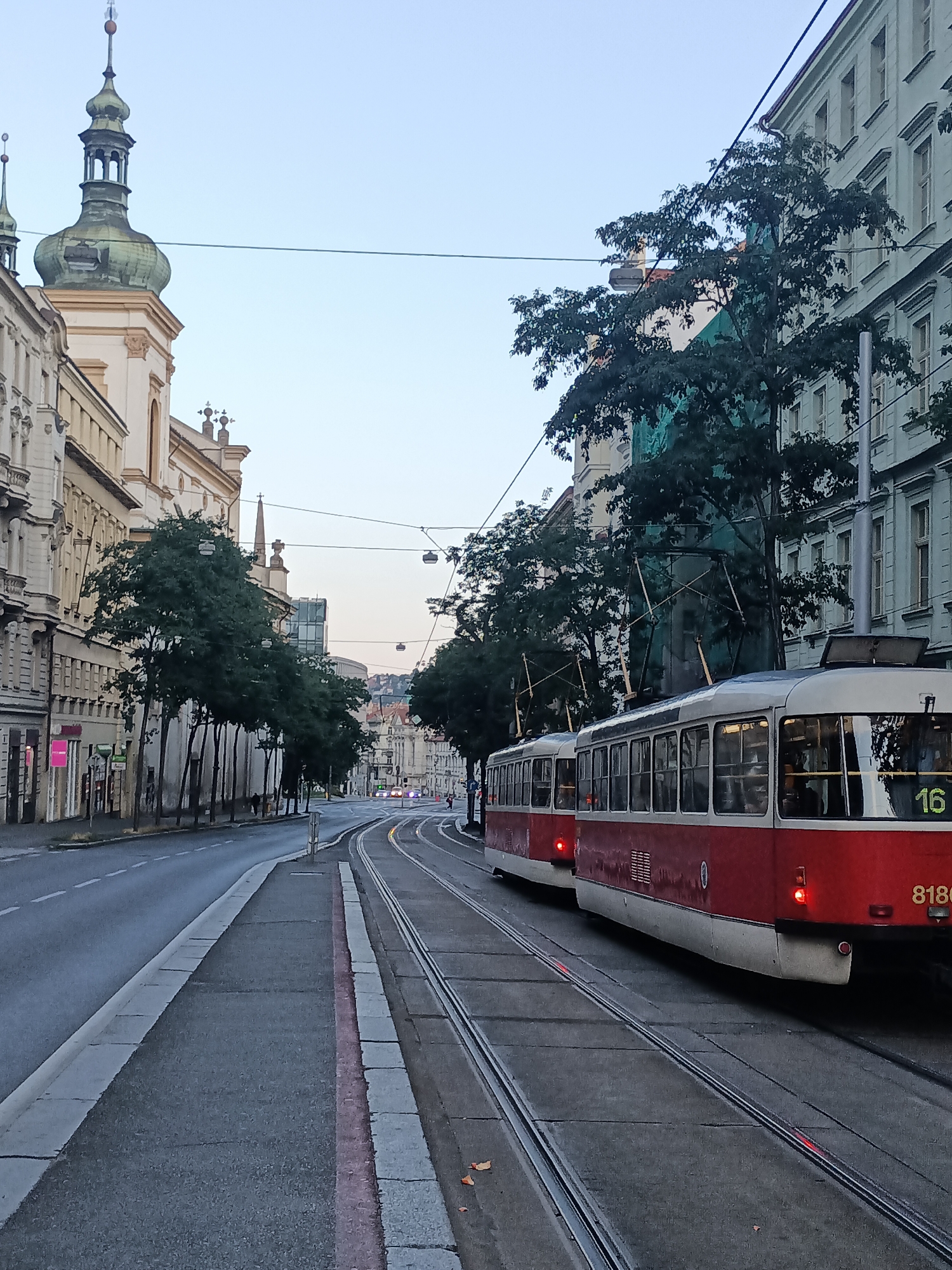 The image captures a view of a street in Prague, Czechia, likely in the Vinohrady district, characterized by its tram infrastructure and classic European architecture. In the foreground, two red and white trams are parked on parallel tracks, with one clearly displaying the number "16" and "8180" on its side. The trams appear to be stationary. The street is paved, with tram tracks running down the center. On the left side of the street, a sidewalk is bordered by a row of trees. Beyond the sidewalk, a building with an ornate facade and a distinctive green-domed tower is visible, suggesting a historical or religious structure. To the right of the street, a light-colored building with multiple windows lines the pavement. The sky above is a clear, pale blue, indicating either early morning or late evening. The scene is largely devoid of people, with only a few indistinct figures visible in the distance on the sidewalk further down the street. The overall impression is one of a quiet urban setting, possibly during off-peak hours.