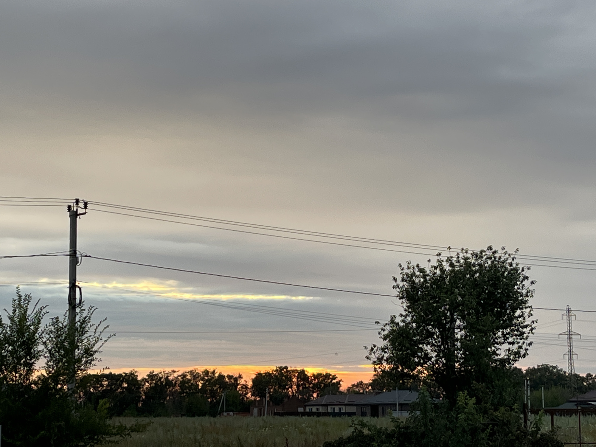 A view of a sunset over a rural landscape. The sky is filled with clouds, with a strip of bright orange and yellow visible near the horizon. A power pole with wires runs across the frame from left to right. In the foreground, there are trees and tall grass. In the midground, there are houses and more trees. In the background, there is a transmission tower. The image appears to be taken from a low angle. The setting is likely a village or outskirts of a city.