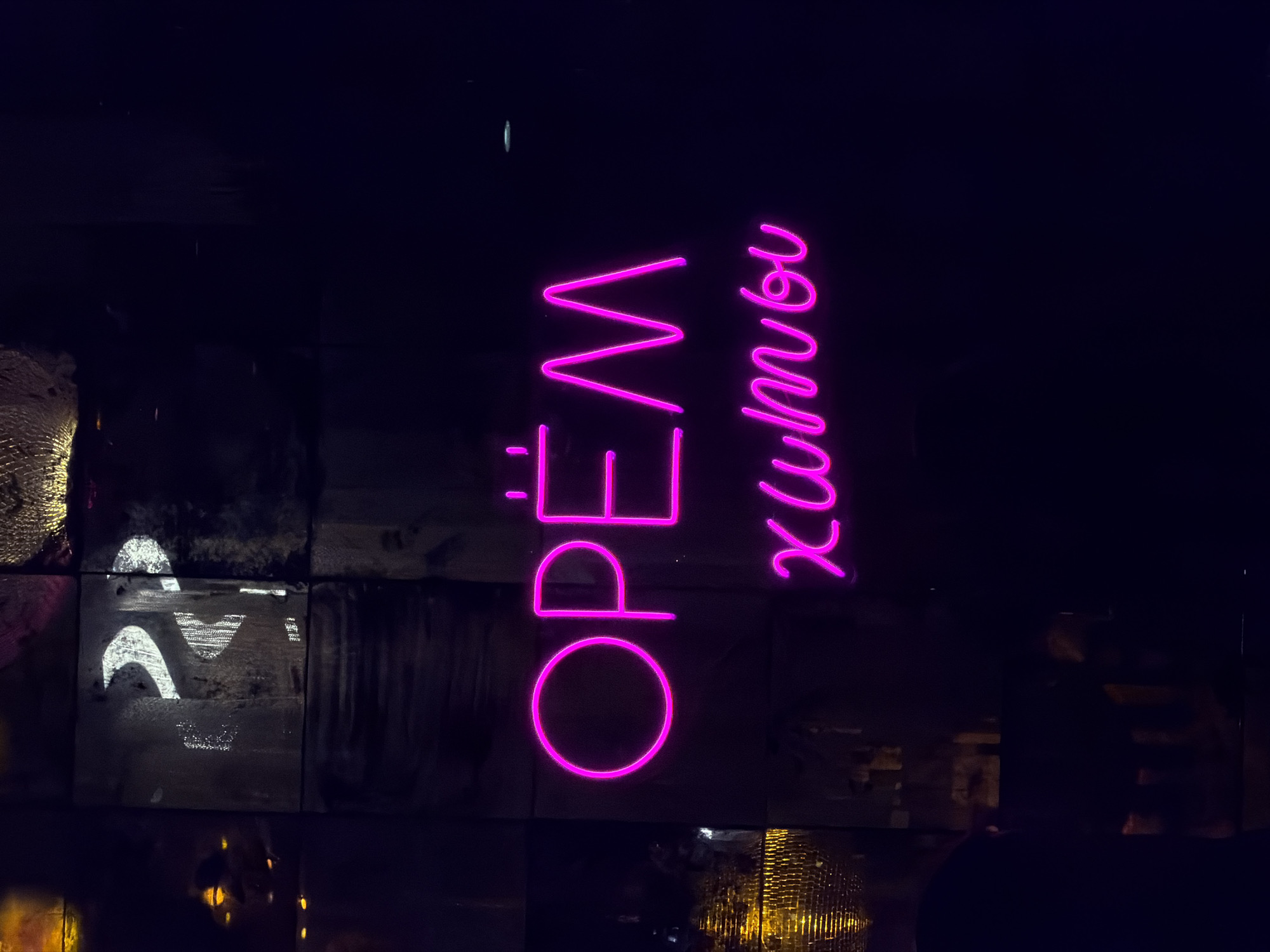 The image shows a neon sign that reads "OPEN ХАМNОИ" in Krasnoyarsk, Russia. The sign is displayed vertically with the word "OPEN" stacked on top of "ХАМNОИ". The letters are illuminated in a bright pink or magenta color. The background is dark and appears to be a wall constructed of various materials, including textured and reflective surfaces. Some of the materials reflect light, indicating other light sources in the vicinity.