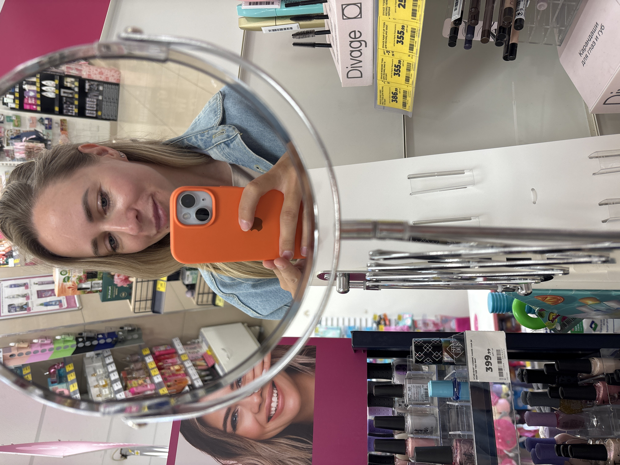 The image shows a woman in a store, reflected in a large, circular mirror. She is holding an orange smartphone up to take a selfie. She is wearing a light blue denim jacket over a light-colored t-shirt and has blonde hair pulled back from her face. Her right hand is visible holding the phone.
In the background, shelves are filled with various cosmetic products, including nail polish bottles and mascara tubes. The store appears to be a beauty supply or cosmetics shop. A large advertisement for makeup is partially visible on the left side of the mirror's reflection, showing a woman's face and smiling mouth.
The reflection also shows aisles of products, with shelving units and price tags. The floor appears to be tiled. The overall environment suggests a retail setting. The location provided is Chaikovsky, Russia, indicating this is a store within that city.