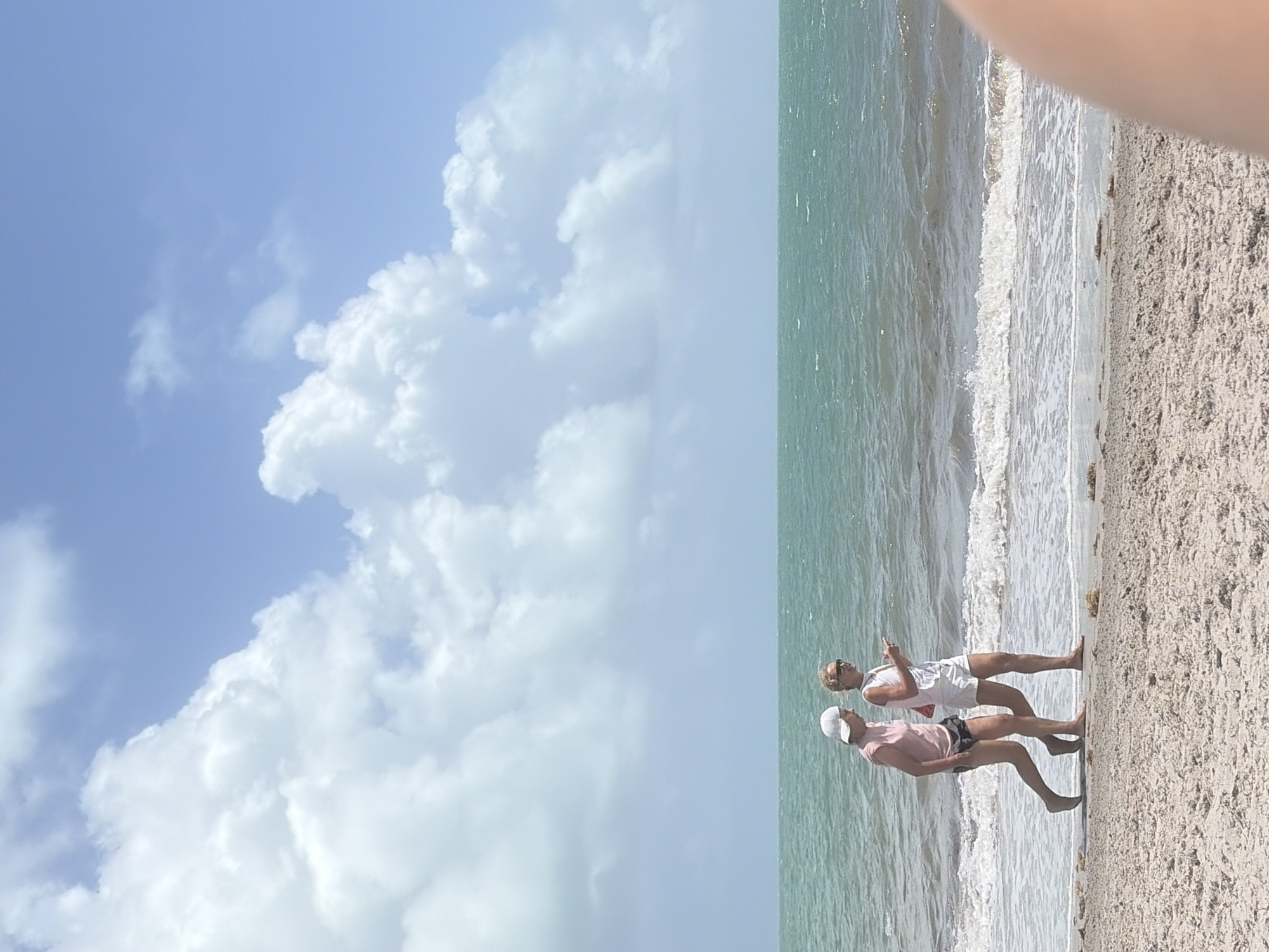 The image depicts two individuals on a beach. The environment includes sand, ocean water, and a sky with clouds. The sky takes up most of the frame with bright blue interspersed among white puffy clouds. The sand and surf run lengthwise with two people in the middle of the frame. One individual is wearing a pink shirt and dark-colored shorts with a white hat. The other individual is wearing a white top and white shorts. It appears the location is a beach in Key Biscayne, Florida, based on the landscape and climate.