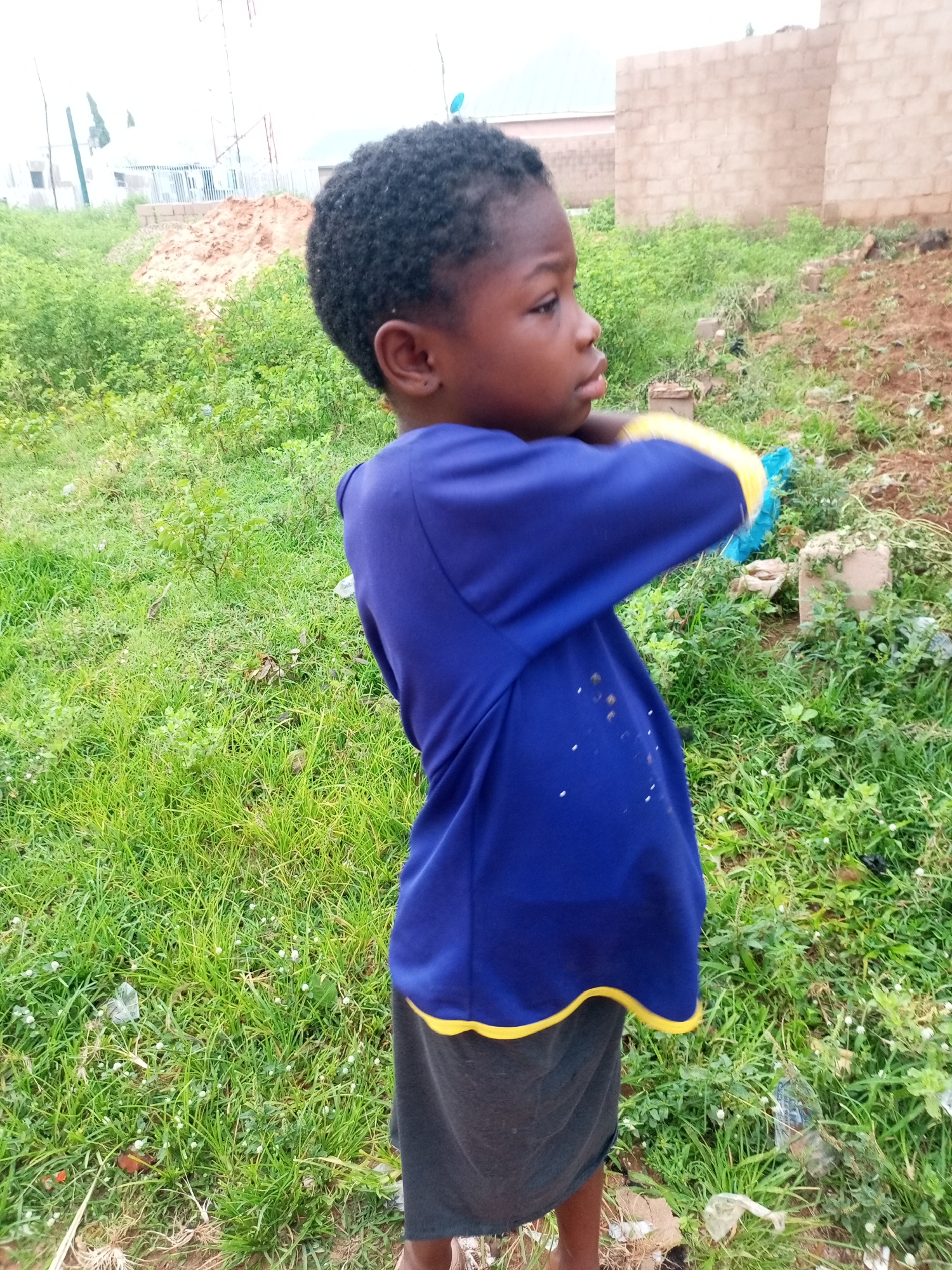 A medium shot captures a young Black child from the chest down, facing left in a grassy area, possibly in Tempure, Nigeria. The child wears a royal blue long-sleeved shirt with yellow trim around the cuffs and hem, and a dark grey skirt. The child's right arm is bent, with their hand near their neck, and they are looking to their right, away from the camera. The background features green grass, scattered debris including plastic bags and bottles, and a dirt mound with some construction materials. Further in the background, there are unfinished brick buildings. The sky is overcast and bright, suggesting daylight.