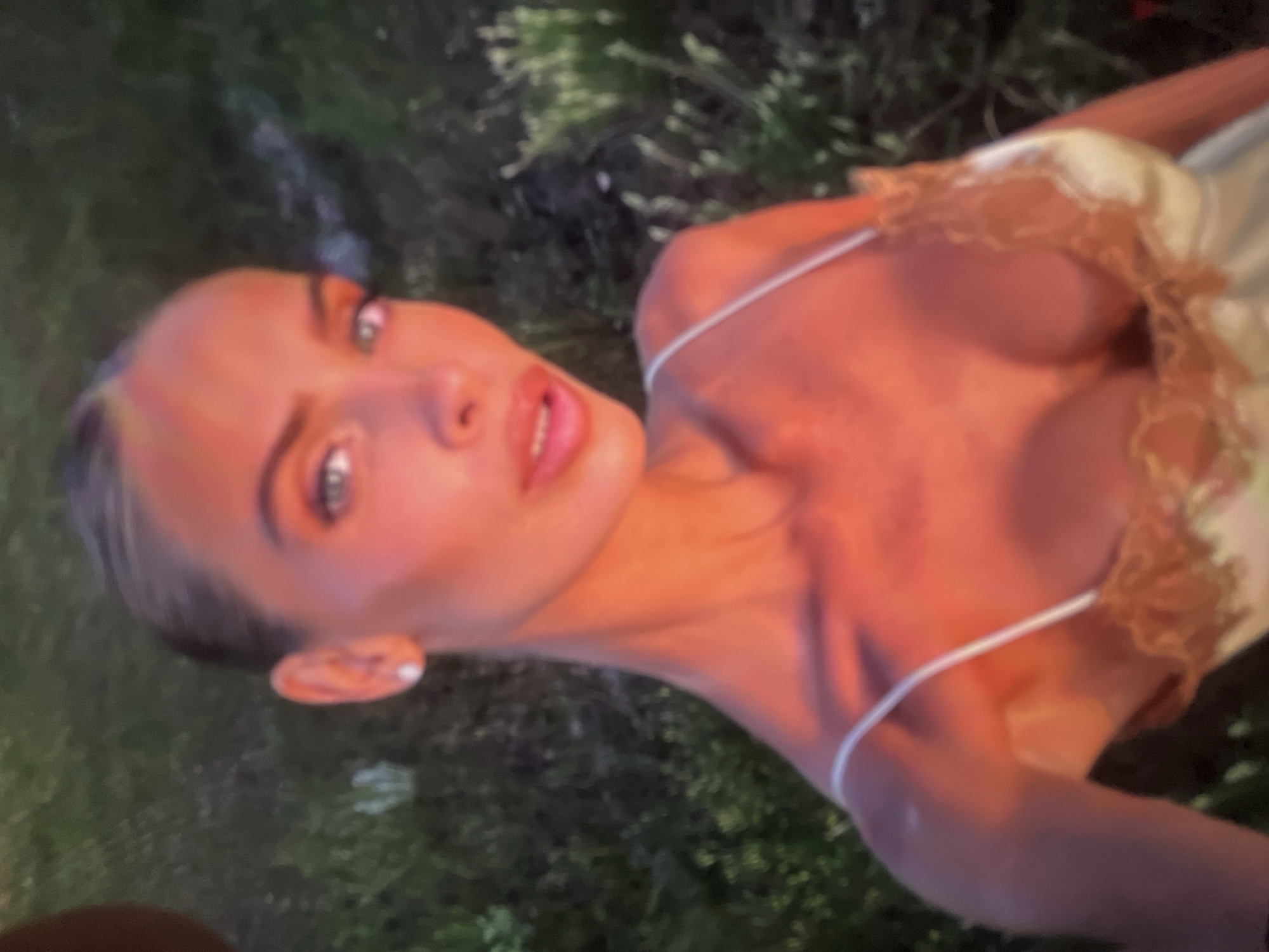 A woman with dark hair pulled back and blue eyes, wearing a white camisole with lace trim, is pictured outdoors in the late afternoon sun. She is looking towards the camera, with her mouth slightly open. The background consists of green foliage. The lighting suggests a warm, possibly sunset, illumination.