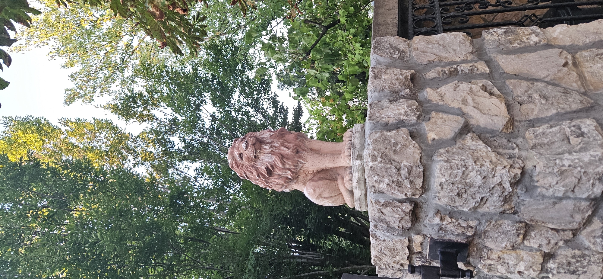 A terra-cotta lion statue sits atop a rough-hewn stone pillar. The statue depicts a lion in a seated position, facing forward with its head tilted slightly upwards. The lion has a prominent mane with textured detailing and appears to be made of clay or concrete. The stone pillar is constructed from irregularly shaped, light-colored stones with visible mortar. Behind the pillar and statue, a dense arrangement of green trees with dappled sunlight filtering through the leaves forms the background. To the right of the pillar, a section of ornate black metal fencing is visible. The scene appears to be outdoors, likely in a garden or park setting. The provided image is a photograph taken in Hévíz, Hungary.