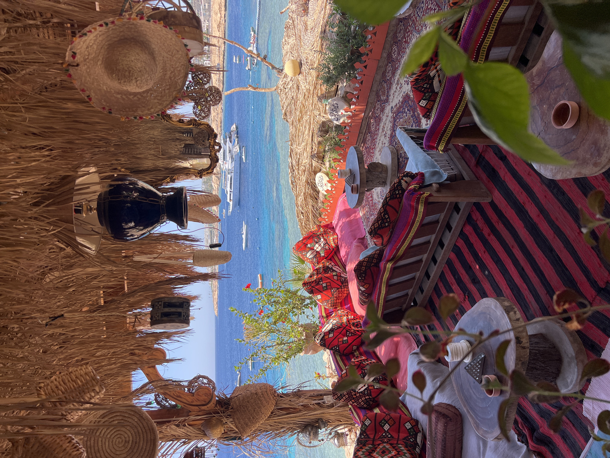 This image depicts an outdoor seating area with a view of the Red Sea, likely in Sharm El Sheikh, Egypt. The seating area is covered by a thatched roof, adorned with various woven baskets, hats, and decorative lamps. Below the roof, comfortable seating is arranged with numerous colorful cushions featuring intricate geometric patterns. Red and black striped rugs cover the floor, and a larger patterned rug is visible beneath the seating.
In the background, the crystal-clear turquoise waters of the Red Sea are visible, dotted with several boats and yachts. The coastline features buildings and lush greenery. The overall atmosphere suggests a relaxed, bohemian-style setting, possibly a cafe or lounge area overlooking the sea. The natural light and vibrant colors contribute to a sense of tranquility and vacation.