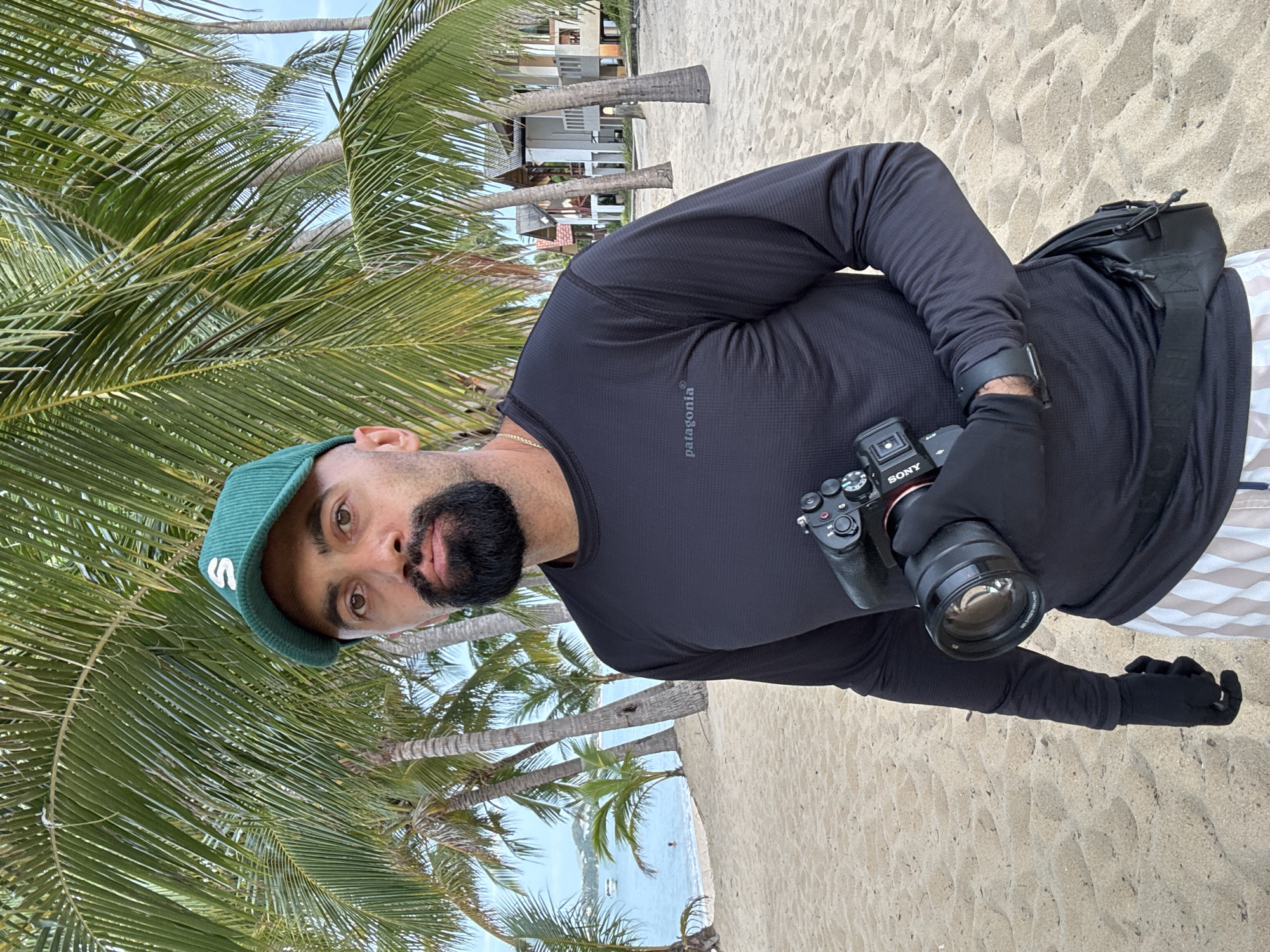 A man with a beard and a green baseball cap stands on a sandy beach, holding a Sony camera with a lens. He is wearing a black long-sleeved shirt with a Patagonia logo and striped shorts. Palm trees and buildings are visible in the background, suggesting a tropical resort location in Ko Samui District, Thailand. The ocean is to the left of the man, with a boat and people in the water.