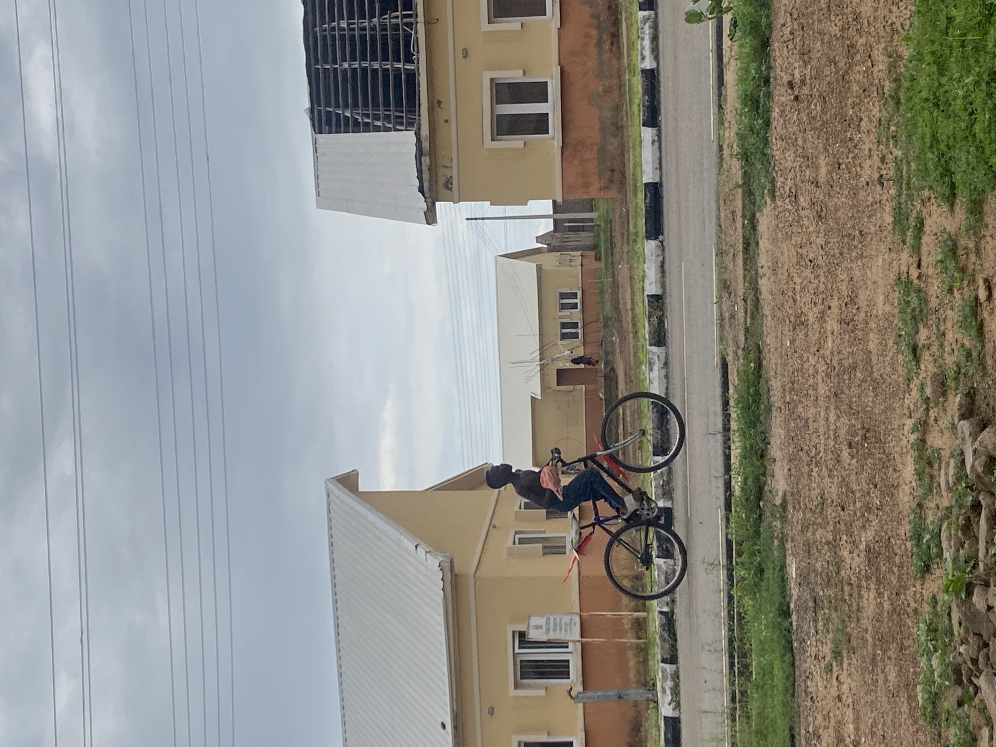 A young Black male is riding a bicycle on a paved road in Bulamare, Nigeria. He is wearing dark blue pants and is shirtless, holding a light pink fabric in his left hand. The bicycle has a red frame and black tires. The scene is set against a backdrop of yellow houses with white roofs, some of which appear to be under construction or have damaged roofs. Power lines are visible in the overcast sky. The road is bordered by a curb with black and white stripes, and the foreground features a patch of dry, brown earth with scattered rocks and some green vegetation.