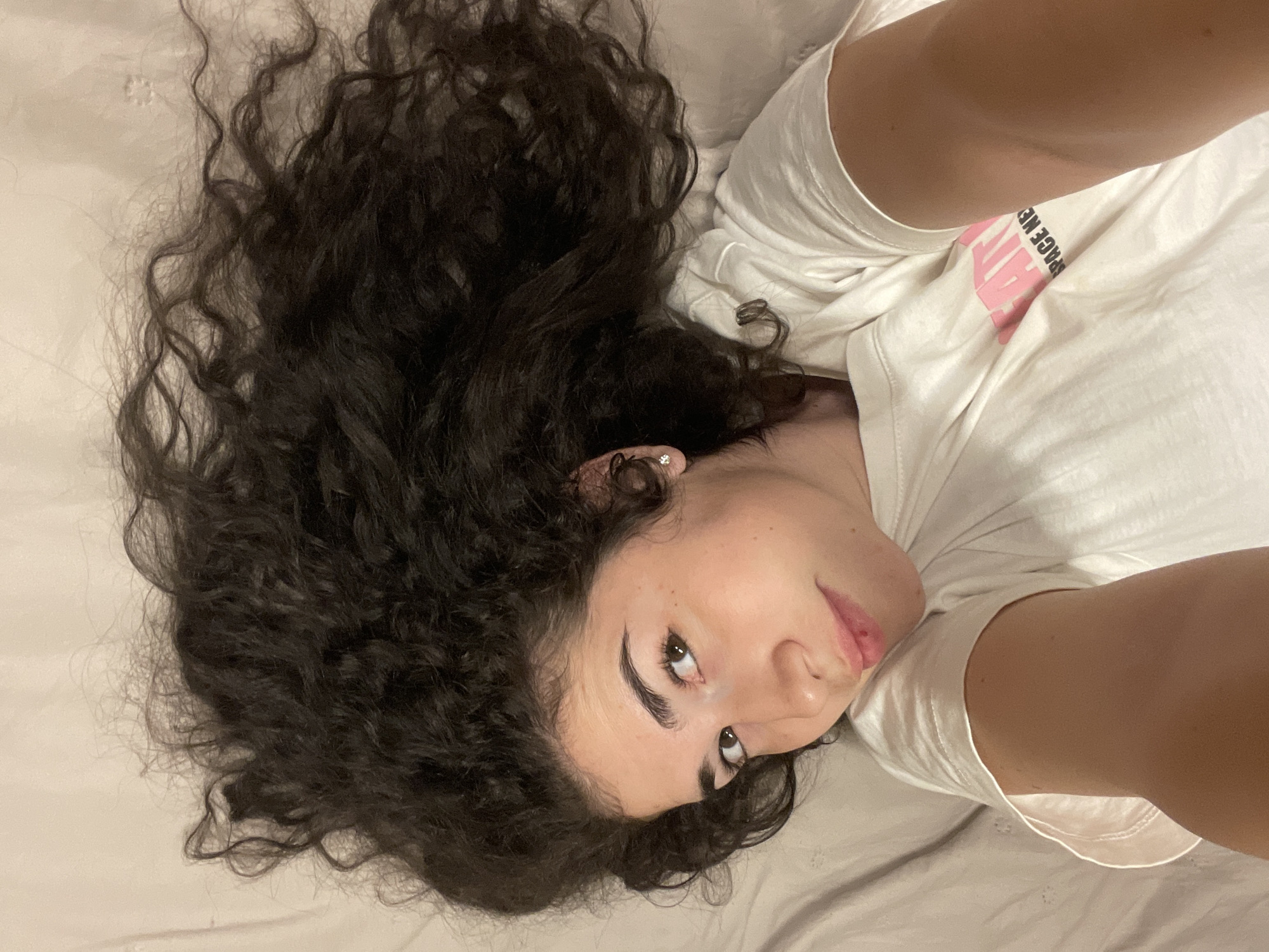 A young person, likely female, is captured in a close-up, high-angle selfie while lying on their back. Their head is turned slightly, and their dark eyes are gazing upwards and to the side, directly at the camera. A subtle, relaxed smile plays on their lips, conveying a casual and perhaps pensive mood.
The individual has long, voluminous dark curly hair that spreads out around their head on a light-colored, rumpled surface, possibly a bedsheet. They are wearing a simple white t-shirt, which features partially visible pink text reading "SPACE KEY". A small, understated earring is visible in their left ear. The lighting is soft and even, suggesting an indoor setting, likely under artificial light or during the evening. The person's arm is visible in the foreground, holding the camera, reinforcing the selfie nature of the shot. This image was taken in Sofia, Bulgaria.