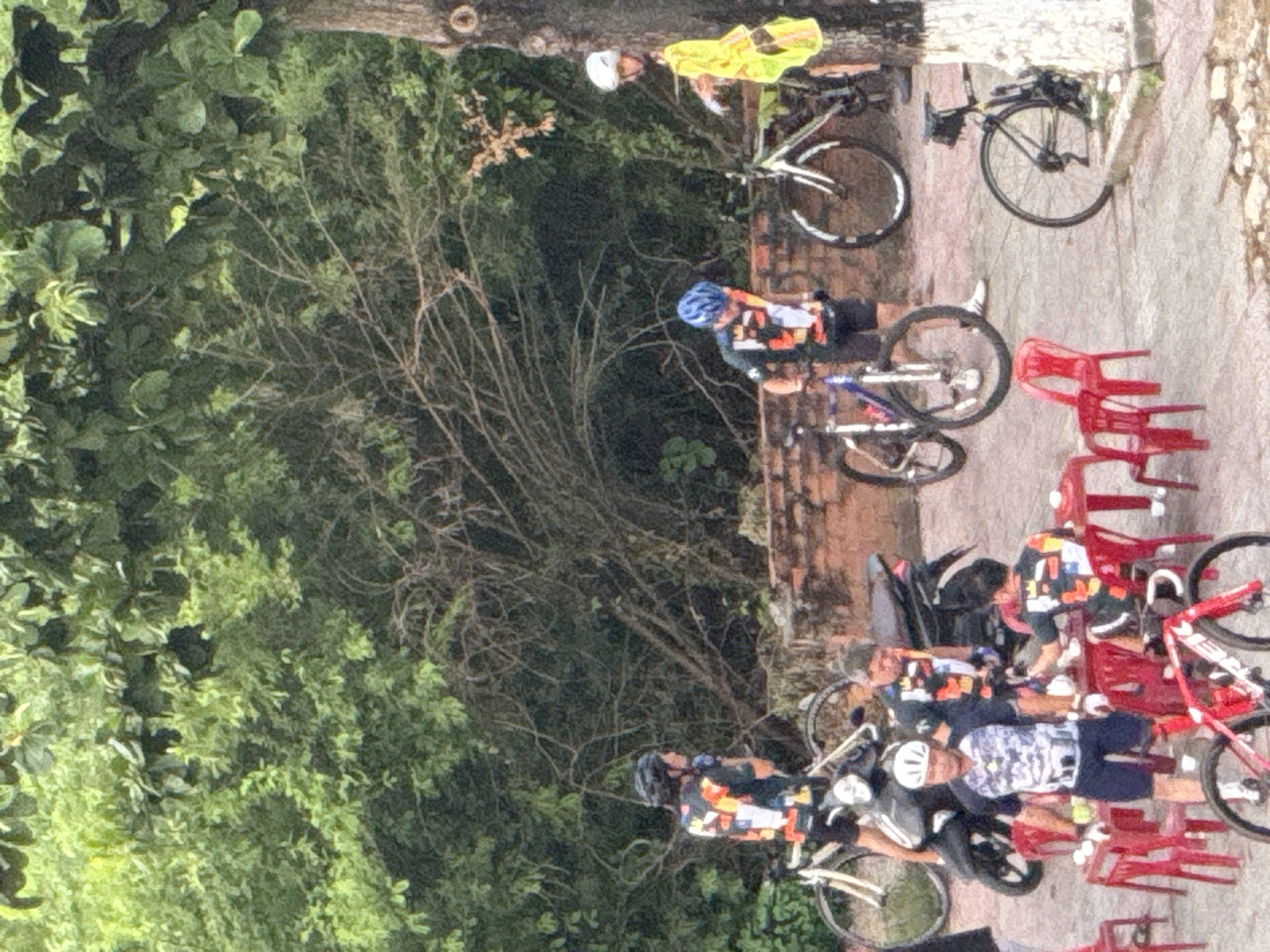 The image shows a group of people, likely cyclists, gathered outdoors. In the foreground, a red bicycle is partially visible. Red plastic chairs are arranged around small tables, some of which hold items like cups. Several individuals are wearing cycling attire and helmets. One person is wearing a bright yellow jacket. Bicycles, including one on a stand, are present in the scene. A motorcycle is also visible behind some of the people. The background consists of dense green foliage and trees, with some exposed brickwork and tree trunks. The lighting suggests it might be daytime, possibly overcast.