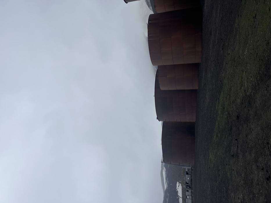 Large, cylindrical, rusty tanks stand in a row against a gray, overcast sky. The tanks appear weathered, with visible paneling. The ground is dark and barren, contrasting with the pale sky. In the distance, hints of snow-capped mountains and a small structure are visible, suggesting a remote or industrial location.