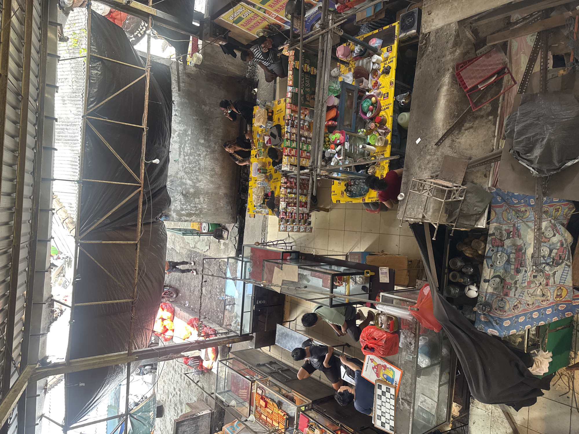 The image captures an overhead view of a bustling outdoor market or food court in Denpasar, Indonesia. The scene is a vibrant mix of commerce and social gathering, with numerous stalls and makeshift dining areas. In the foreground, a few individuals are seated at a stall with glass display cases, one person is looking at a chess board. Further back, several tables covered with yellow tablecloths are occupied by people eating and socializing. These tables are laden with food items, bottles, and packets of snacks, suggesting a food vendor area. Other stalls display various goods, including what appear to be snacks and packaged foods on shelves. The market is covered by a corrugated metal roof and black tarpaulins, providing shade. The ground appears to be paved with concrete tiles. In the background, some produce such as what looks like oranges or mangoes are piled up, and a motorcycle is parked nearby. The overall impression is of a lively, open-air marketplace in an urban setting.