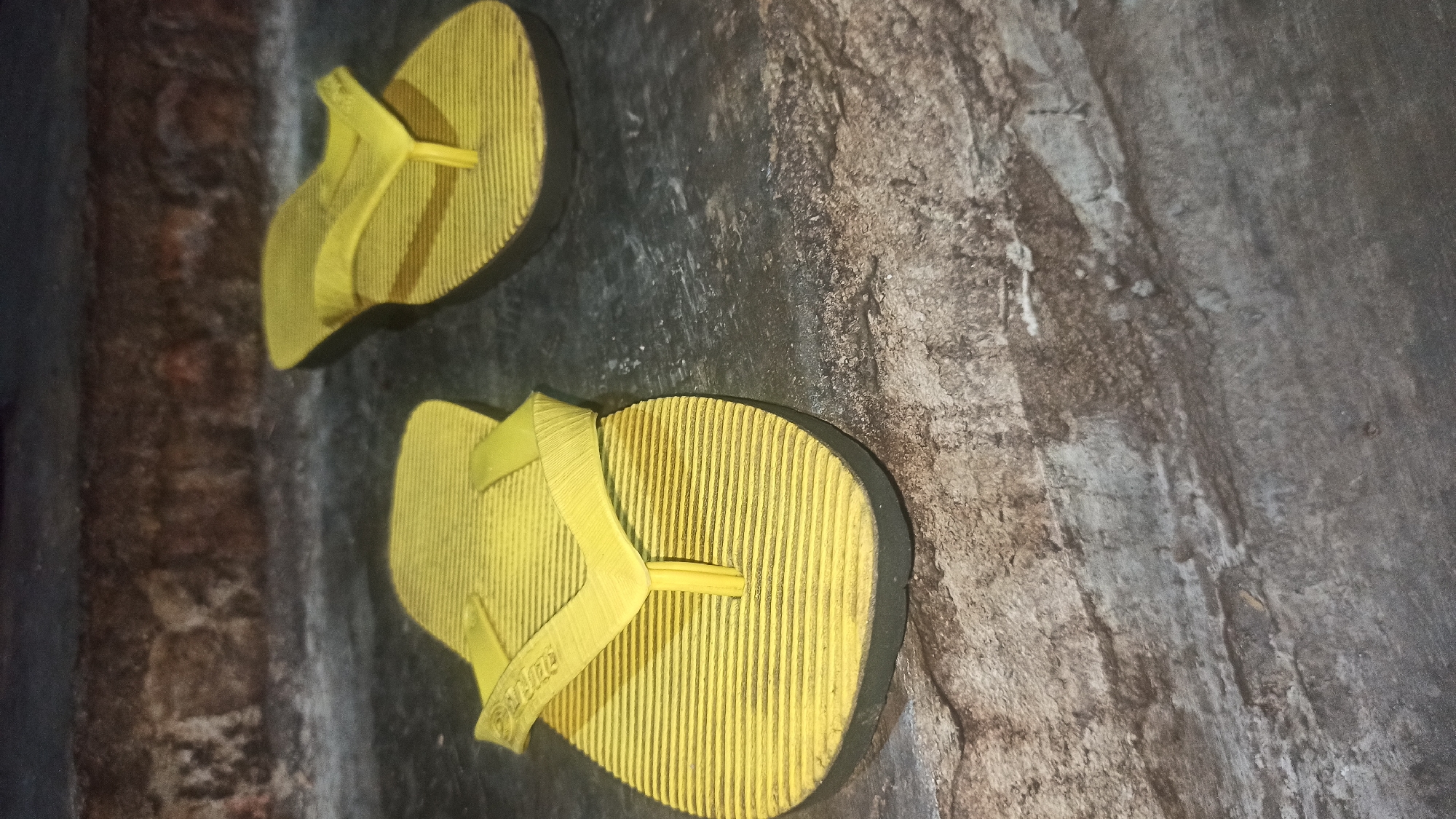This is a close-up, low-angle shot of two yellow flip-flops on a textured, grayish-brown surface that appears to be concrete or stone. The flip-flops have a ribbed texture on their soles and a Y-shaped strap. The one in the foreground is slightly to the left of center, and the other is further back and to the right. The background shows a rough, brick-like texture. There are no people or animals present, and no specific identifiable landmarks of Tuba, Indonesia are visible. The lighting suggests an indoor or shaded outdoor environment.
